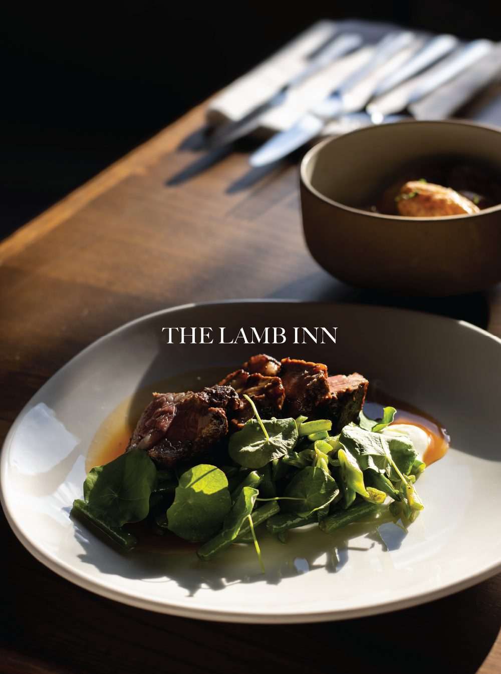 A white plate with a serving of roasted beef with sauce, accompanied by greens. In the background, a black bowl with potatoes and a wooden table with silverware. The Lamb Inn, Cotswolds.