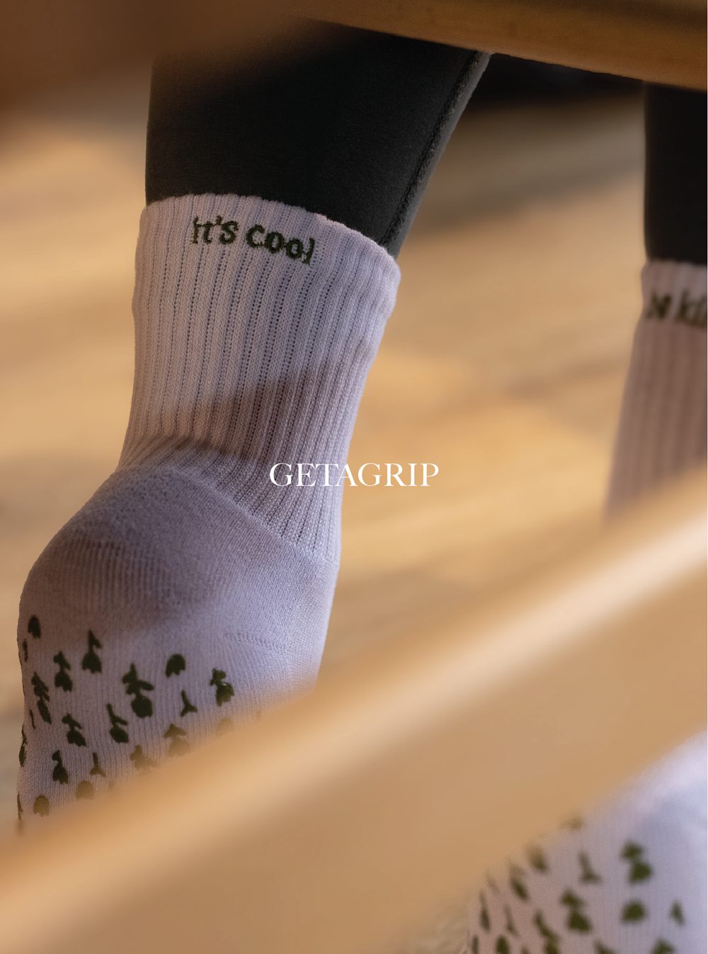 Close-up of a person's foot wearing a white sock with the text 'It's cool' embroidered near the ankle, and a black shoe. The background is blurred with warm tones. Get A Grip Apparel.