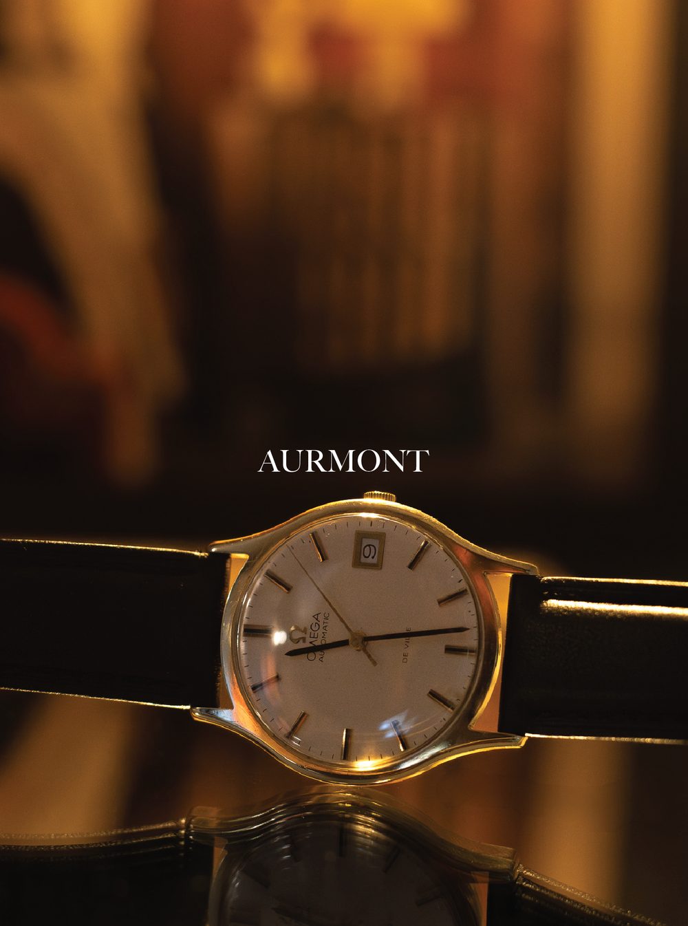 Close-up of a gold Omega De Ville automatic wristwatch with a leather strap on a reflective surface, with a blurred background and the text 'AURMONT' above the watch.