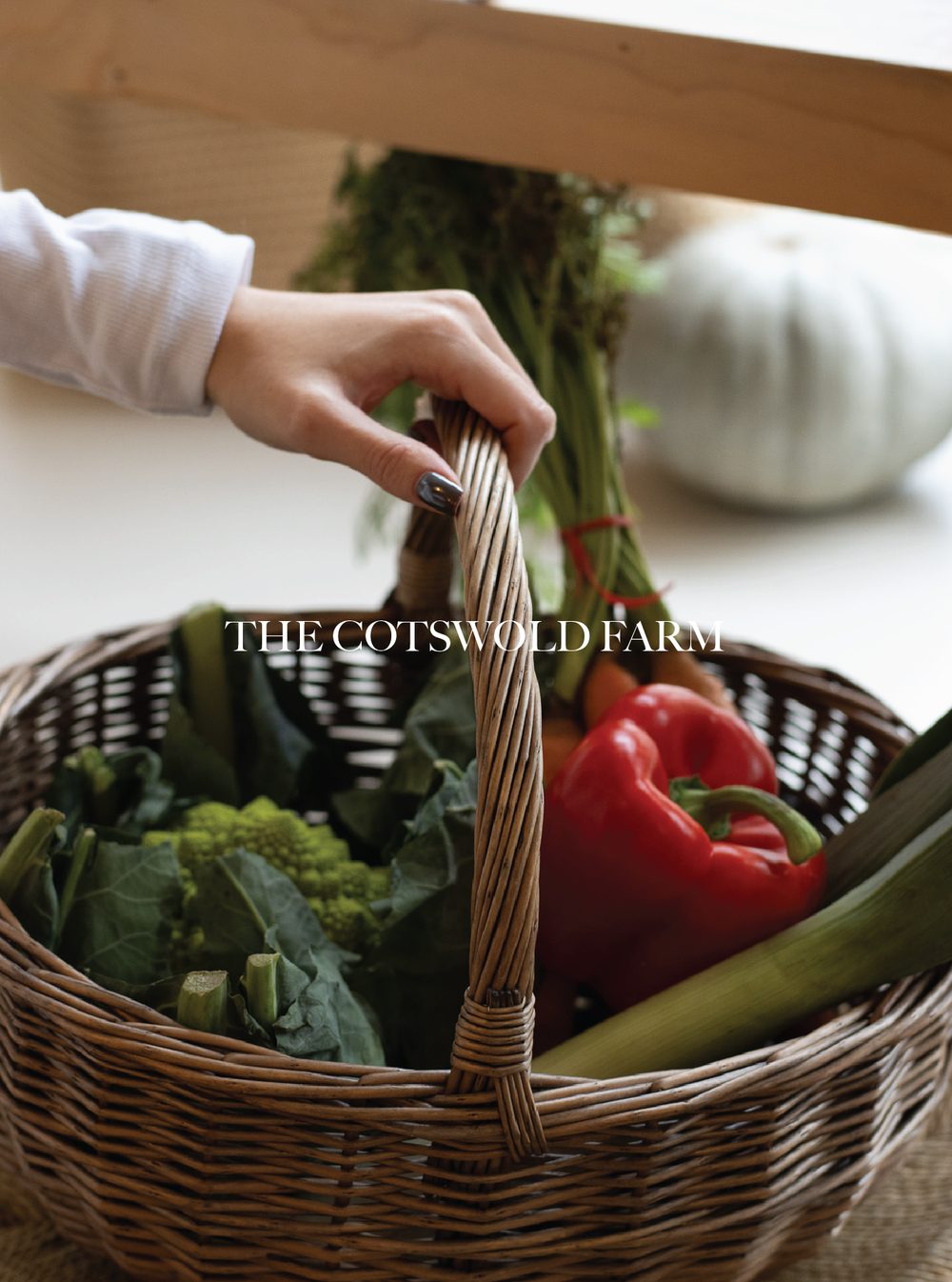 A wicker basket filled with fresh vegetables including red bell peppers, celery, and leafy greens, with a blurred potted plant in the background. The Cotswold Farm.