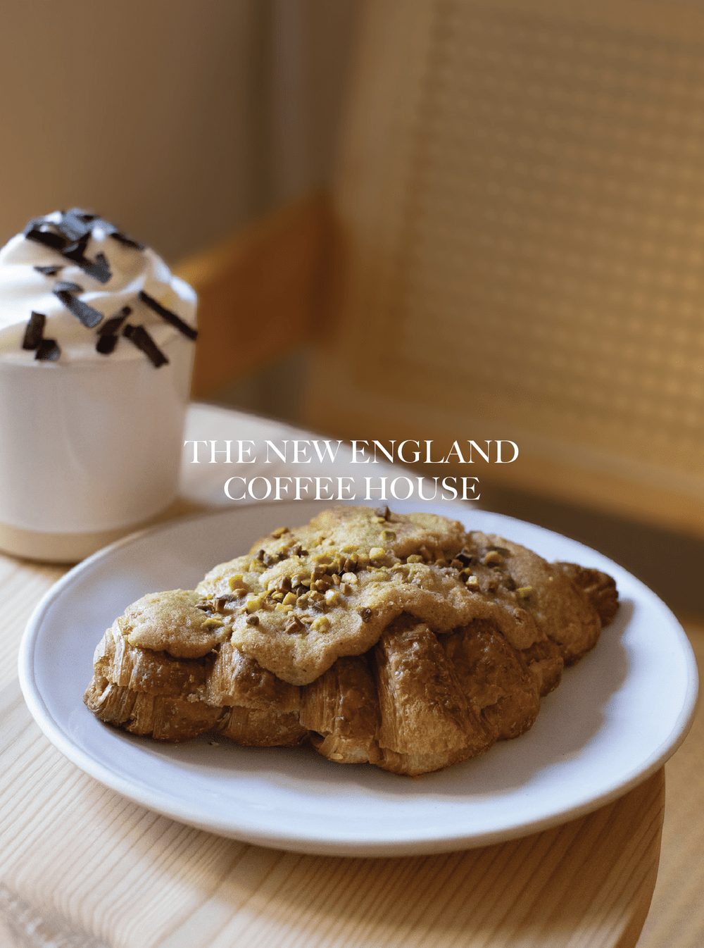 A plate of a croissant topped with a cookie that has pistachio nuts, next to a cup of coffee with whipped cream and chocolate sprinkles, on a wooden surface. The New England Coffee House. Stow on the Wold.