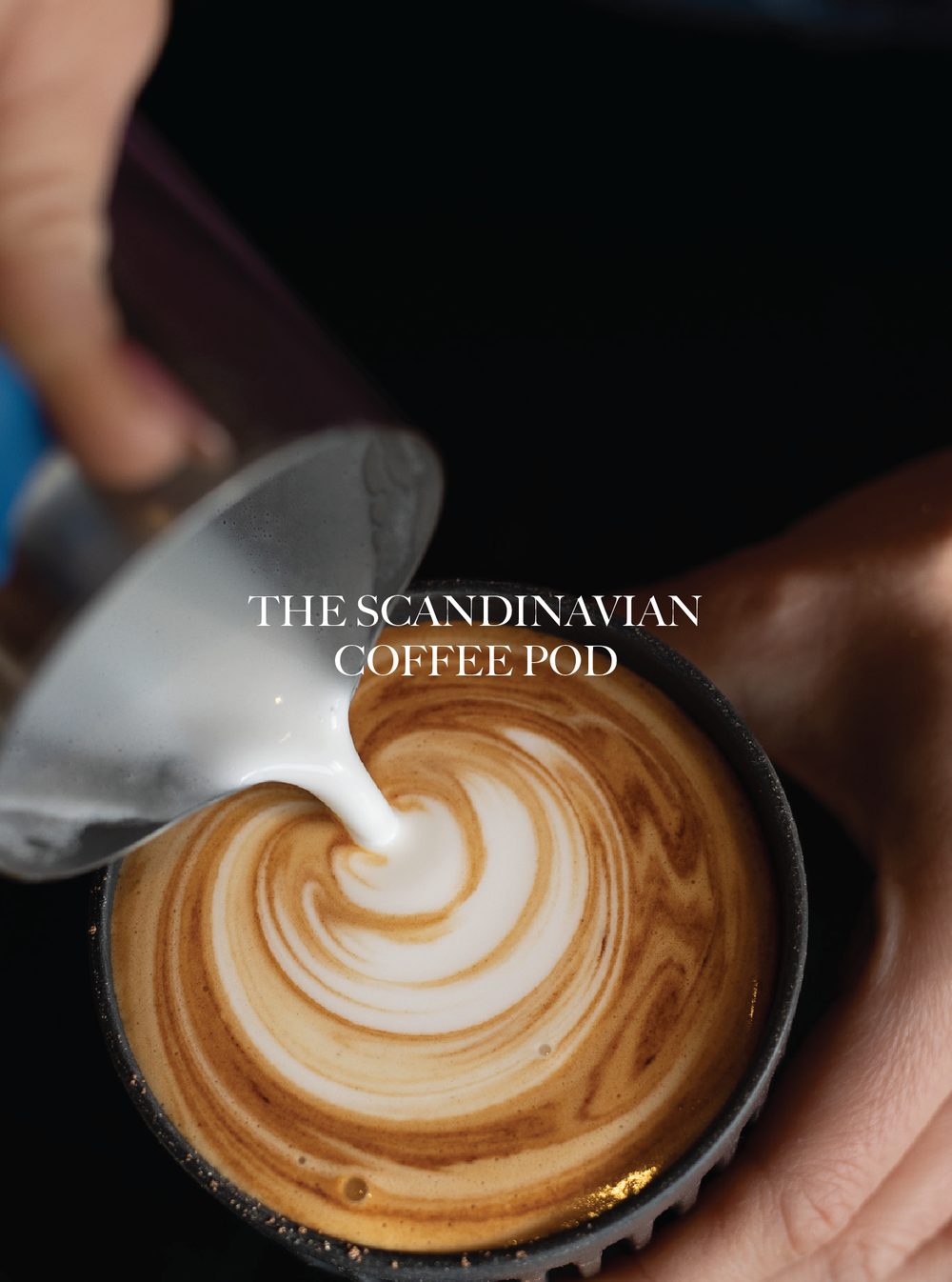 Close-up of a hand pouring steamed milk into a cup of coffee, creating a latte art pattern with a cream swirl design. The Scandinavian Coffee Pod.