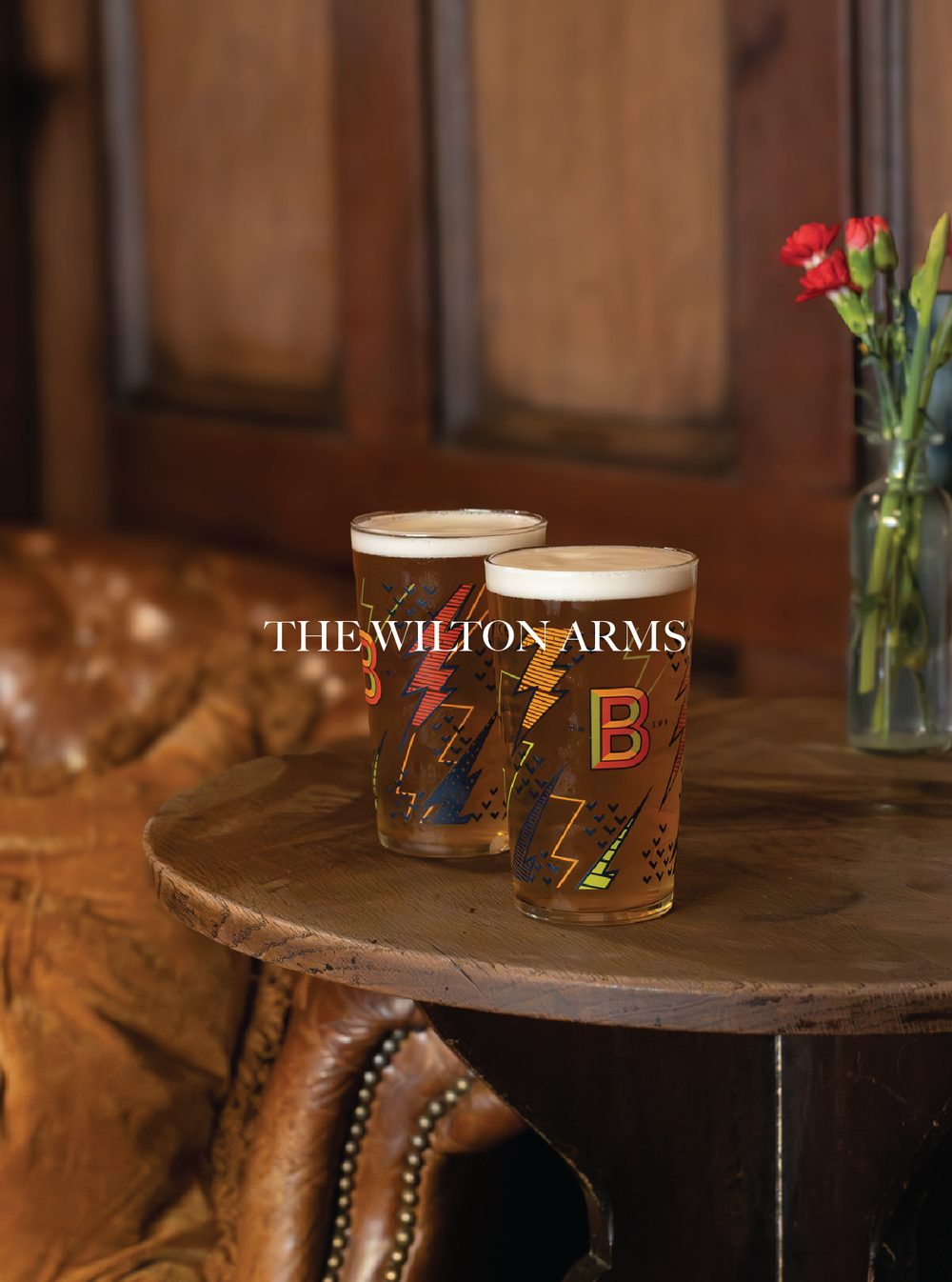 Two pint glasses of beer on a round wooden table, with a bouquet of red flowers in a glass vase in the background. The setting appears to be a cosy bar or pub with wood-paneled walls. The Wilton Arms Pub.