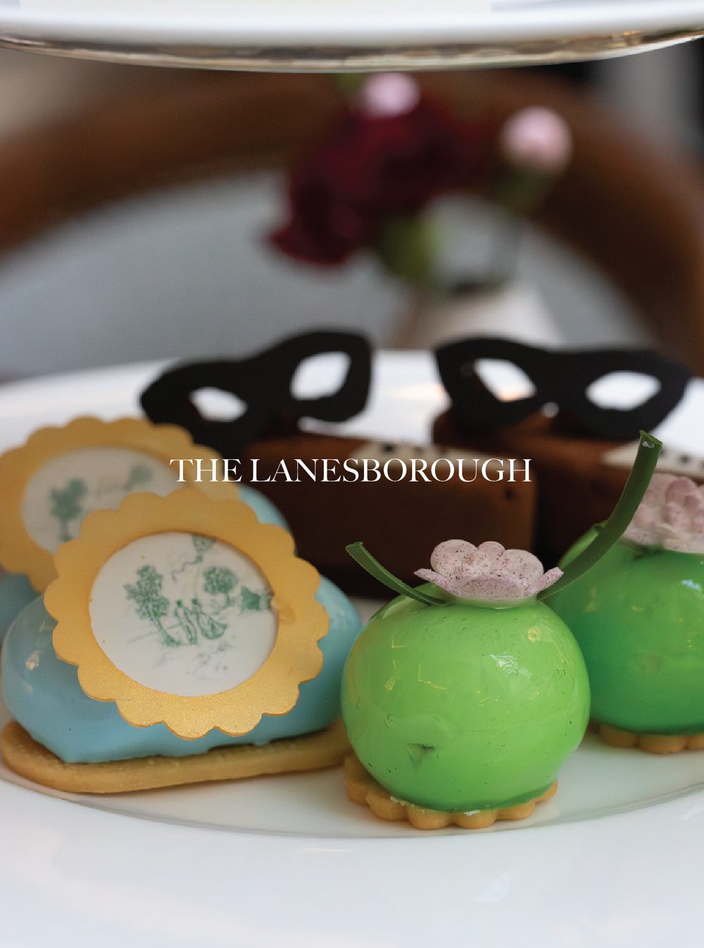 Colorful decorated Easter eggs with one in a bright green color, topped with a small pink and white flower, and another with a vintage picture design and a scalloped yellow border, displayed on a white plate. The Lanesborough.