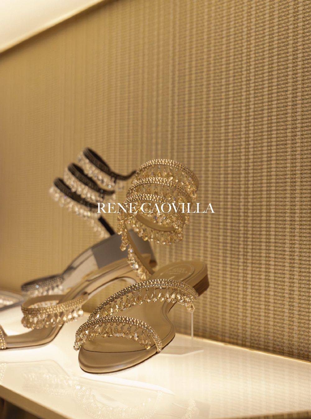 Display of gold and crystal jewelry, including earrings, bracelets, and rings, against a gold textured background. Rene Caovilla.