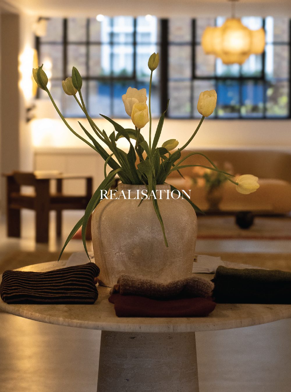A white vase with yellow tulips and green leaves on a round table in a well-lit room with large windows and hanging lights in the background. Realisation Par.