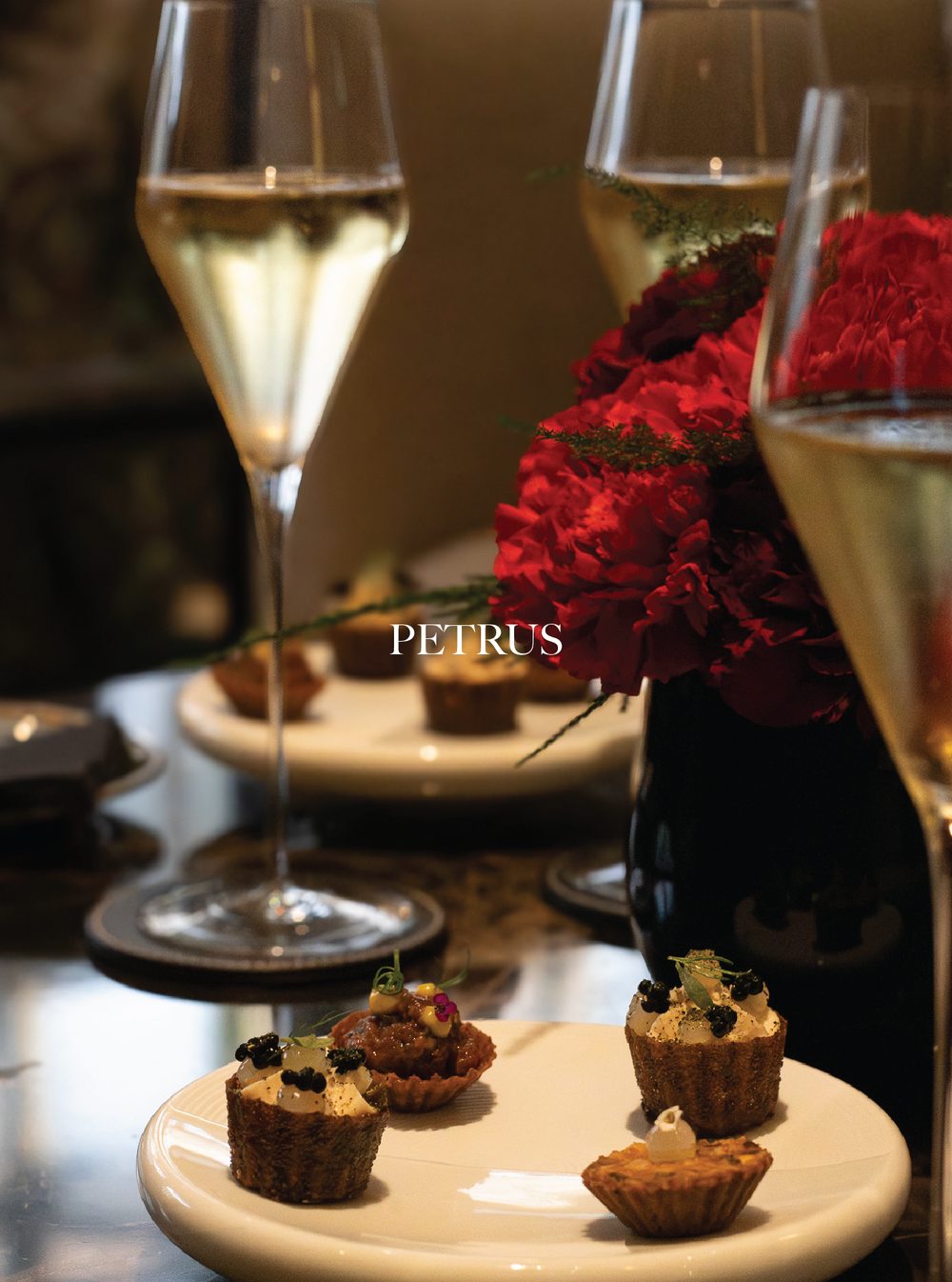 Two glasses of white wine or champagne on a table, with a background of red flowers and desserts on a white plate in the foreground. Petrus. Gordon Ramsey.