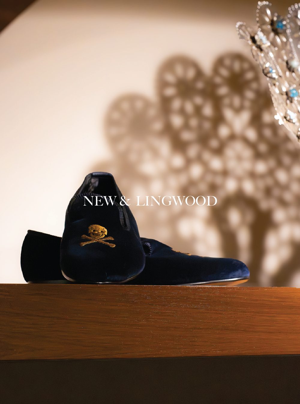 A pair of black shoes with gold embroidered logos on the toes, placed on a wooden surface with a shadow of decorative circular objects and a crown-shaped object in the background, along with the text 'NEW & LINGWOOD'.