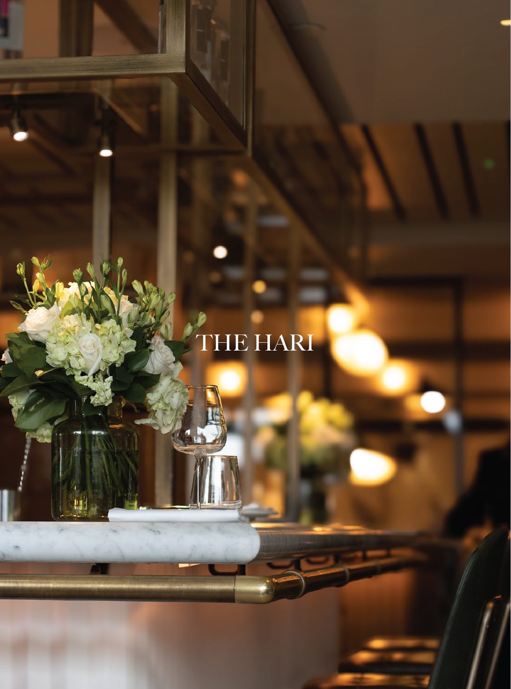 A floral arrangement with white and green flowers, placed on a marble countertop with wine glasses nearby. The background shows warm, dim lighting in a restaurant or event space with modern decor. The Hari, Knightsbridge, Il Pampero.