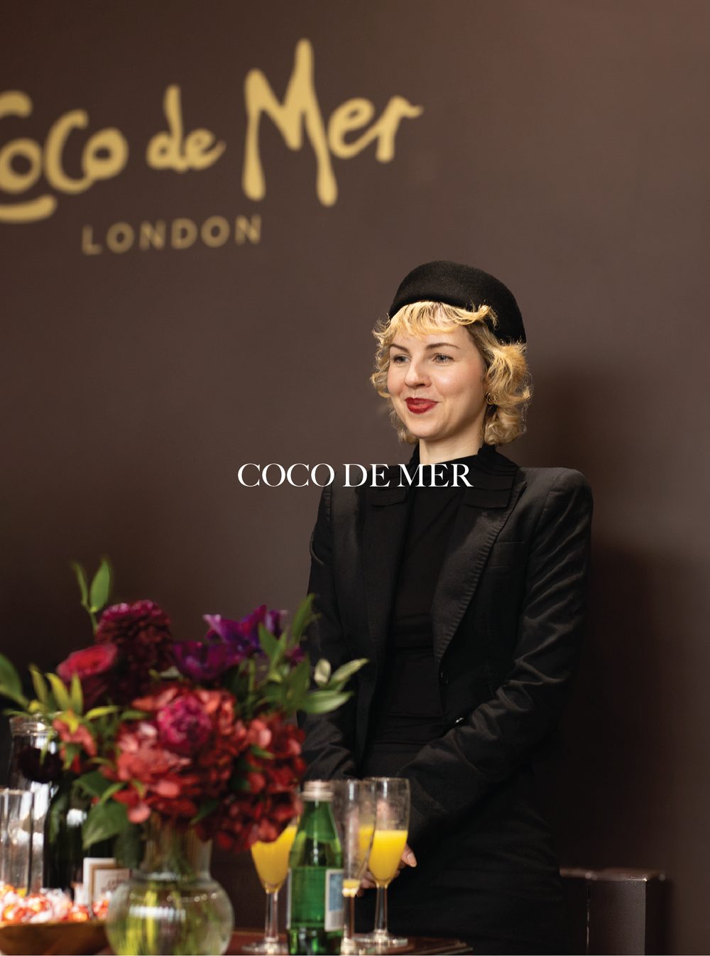 A woman with blonde curly hair wearing a black beret and black blazer standing at a table with flowers and drinks, with a dark background featuring the 'Coco de Mer London' logo.