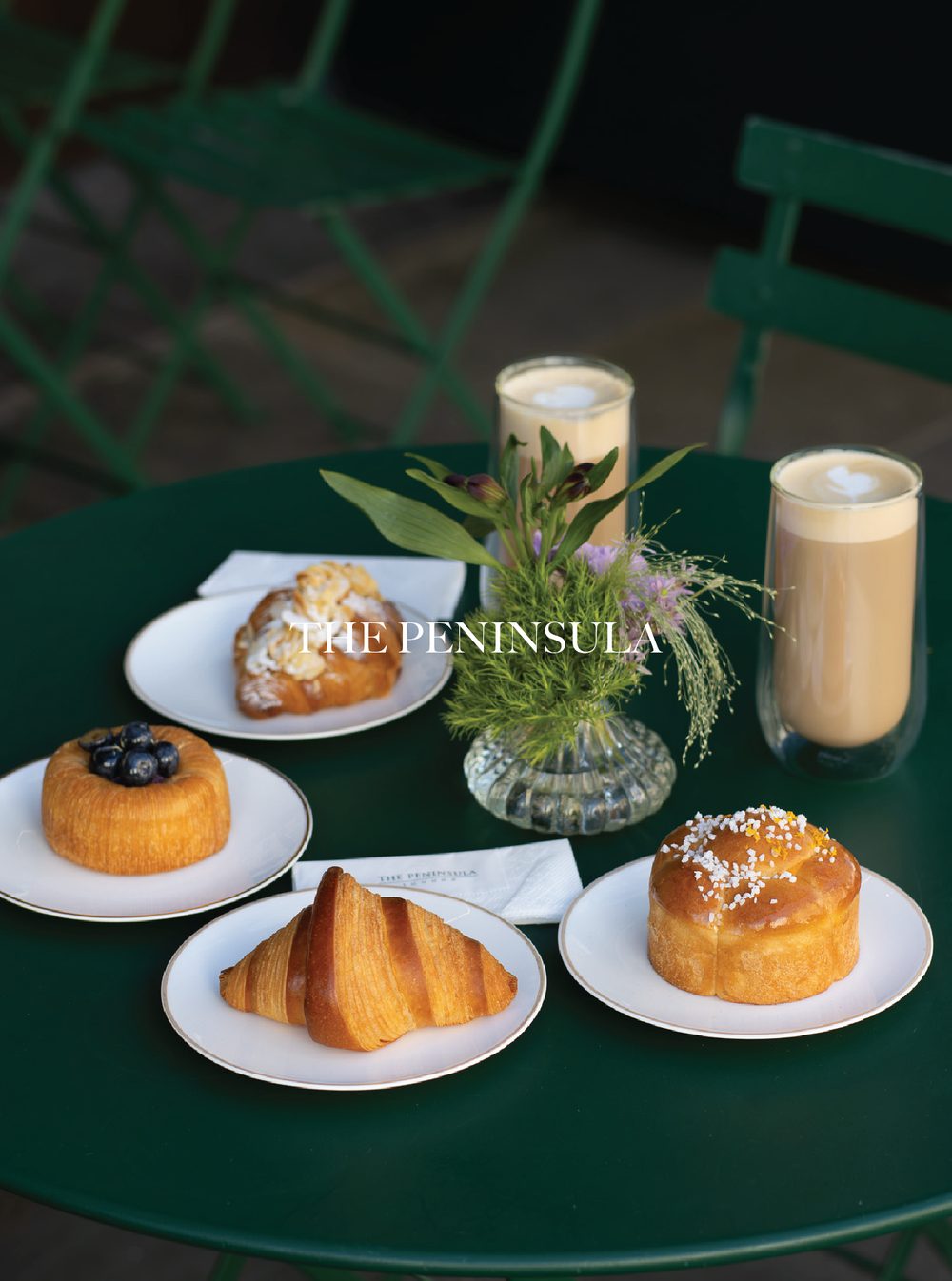 A green table with breakfast pastries, a flower arrangement, and two candles in glass holders. In the background, there are green chairs. The Peninsula.
