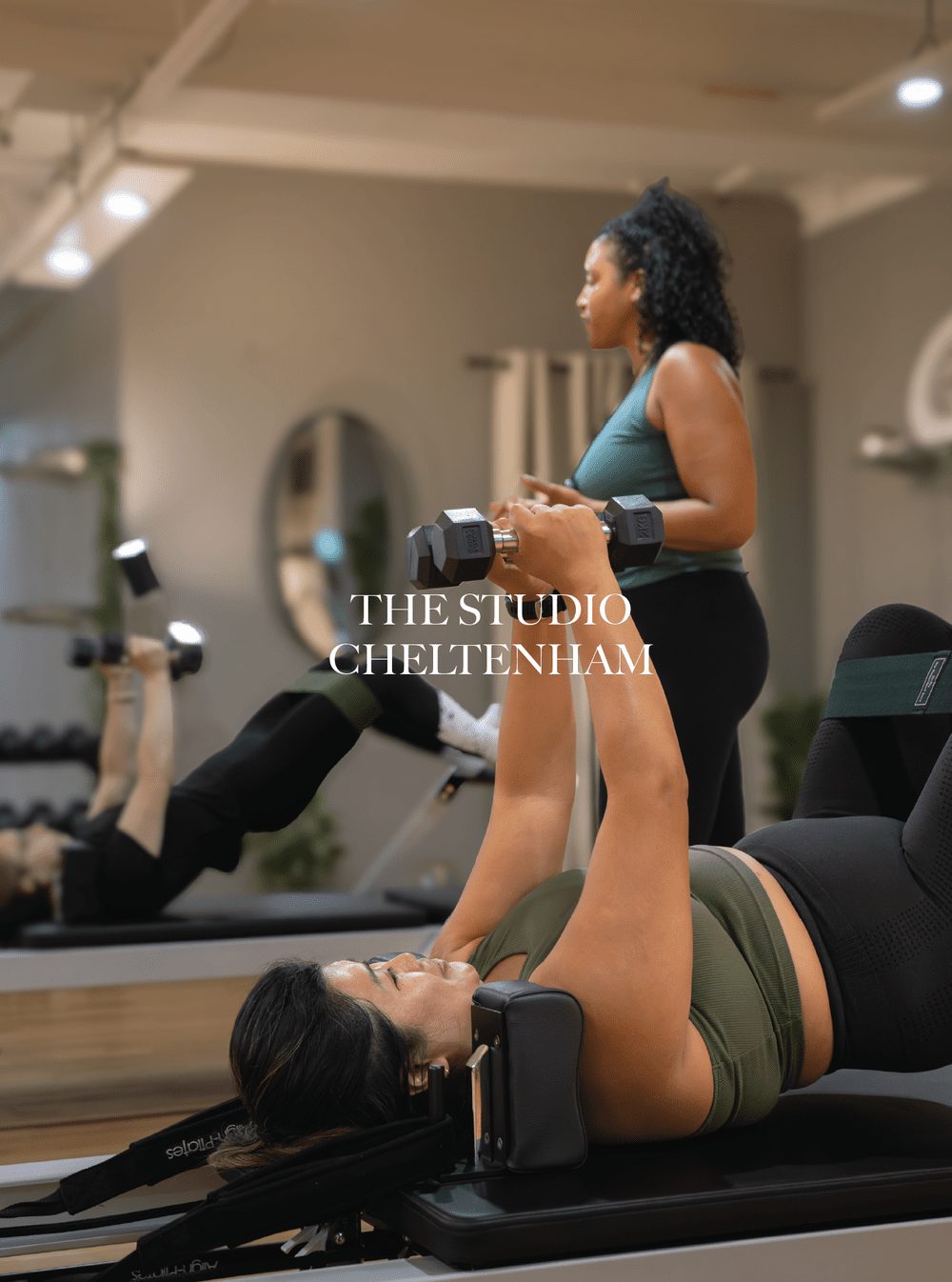 Women exercising on reformer Pilates machines in a fitness studio with a trainer assisting a woman with dumbbells in the background. The Studio Cheltenham.