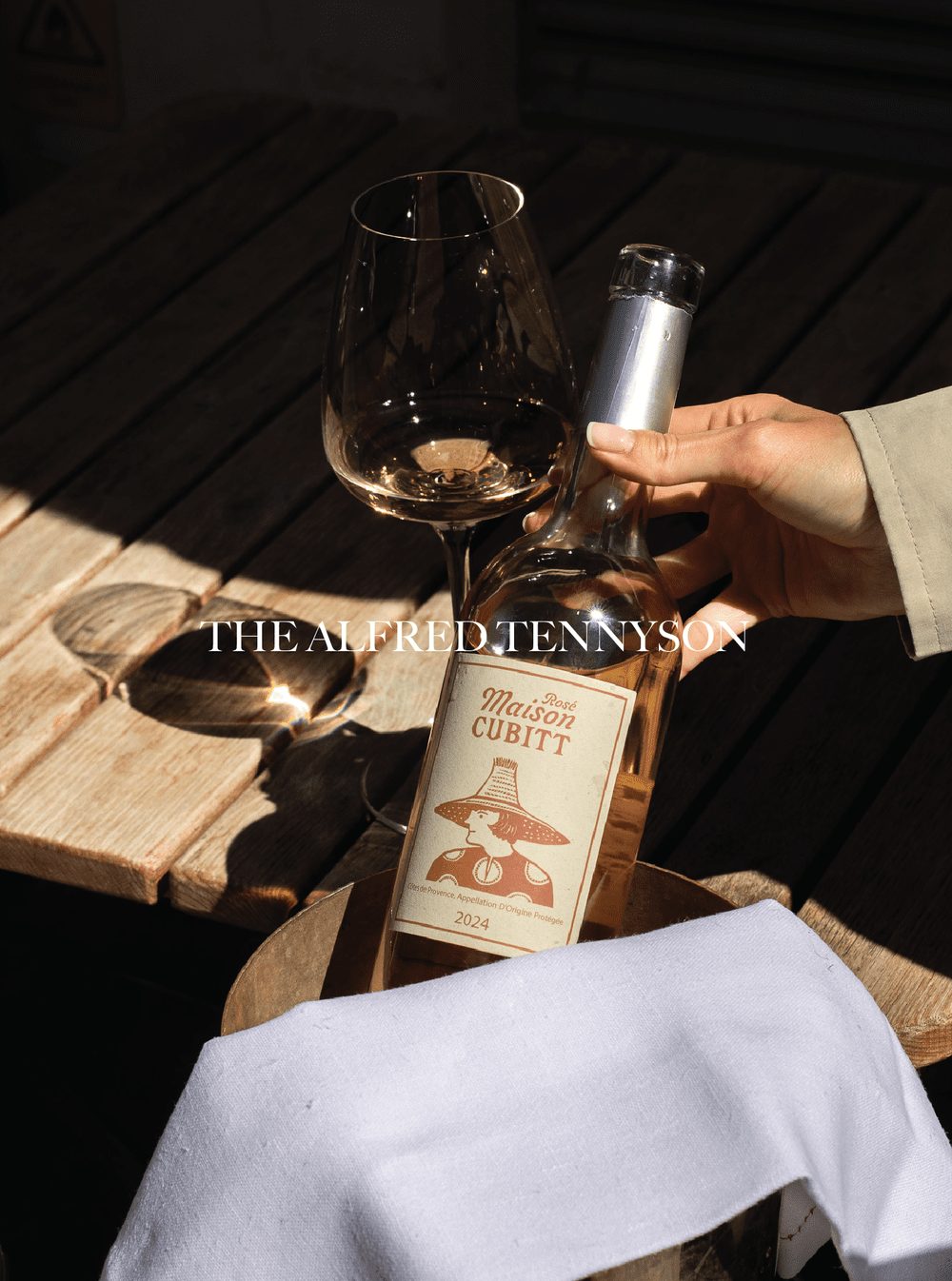 A hand holding a wine bottle labeled 'Maison Cubitt Rosé 2024' next to a glass of white wine on a wooden table with a cloth underneath. The Alfred Tennyson.