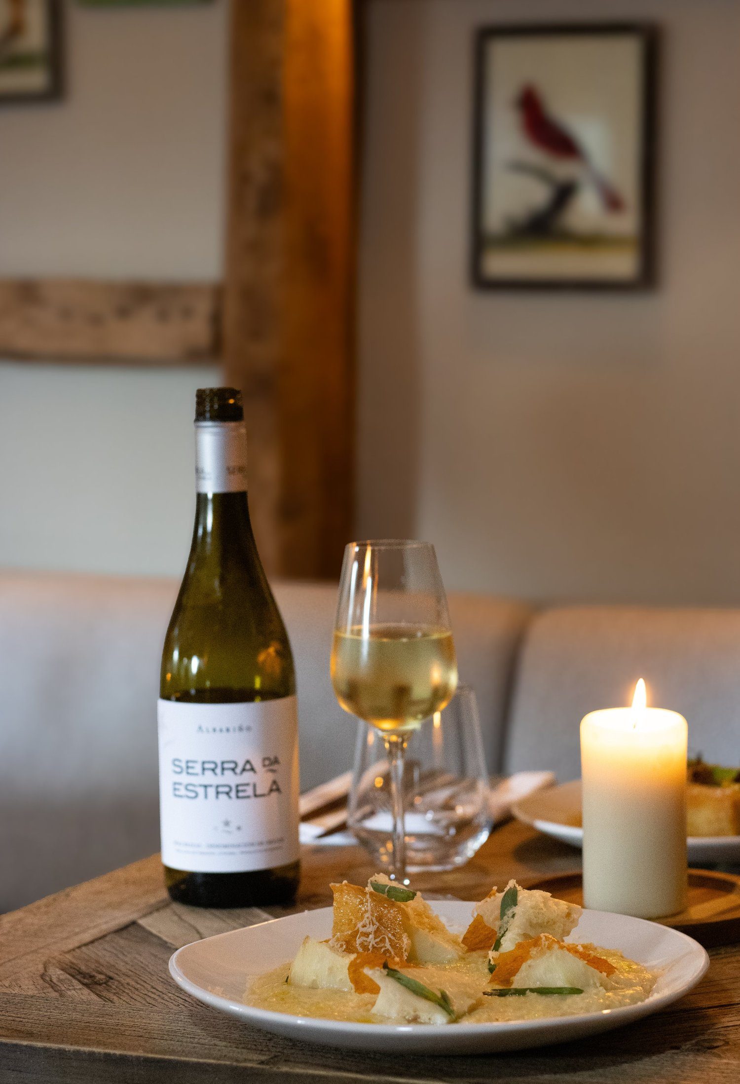 A table setting with a bottle of white wine labeled 'Serra d'Estrela,' a glass of white wine, a lit candle, and a plate of food, in a cozy dining room with framed pictures on the wall. The Lamb Inn.