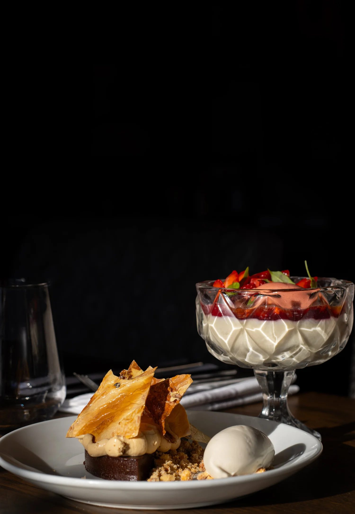 A dessert plate with a chocolate cake topped with cream, caramelized pastry, and white sauce, served with ice cream spheres. In the background, a glass bowl with layered fruit and cream, garnished with fruit slices, is visible against a black backgro