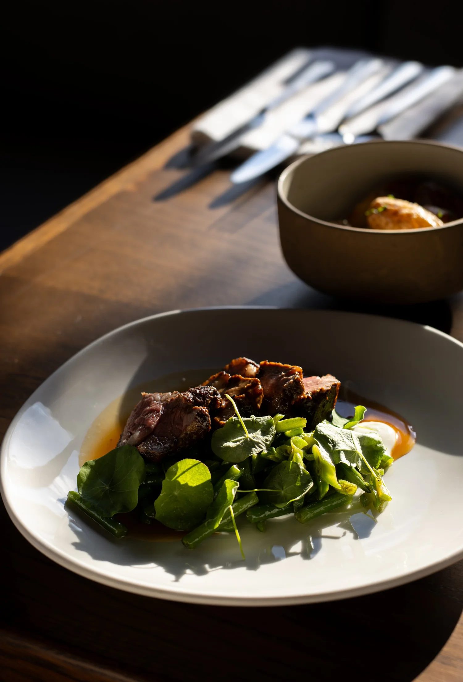 A white plate with sliced meat, green leafy vegetables, and an egg with a runny yolk. In the background, there is a dark bowl with a piece of meat, and a wooden table with metal utensils. The Lamb Inn.