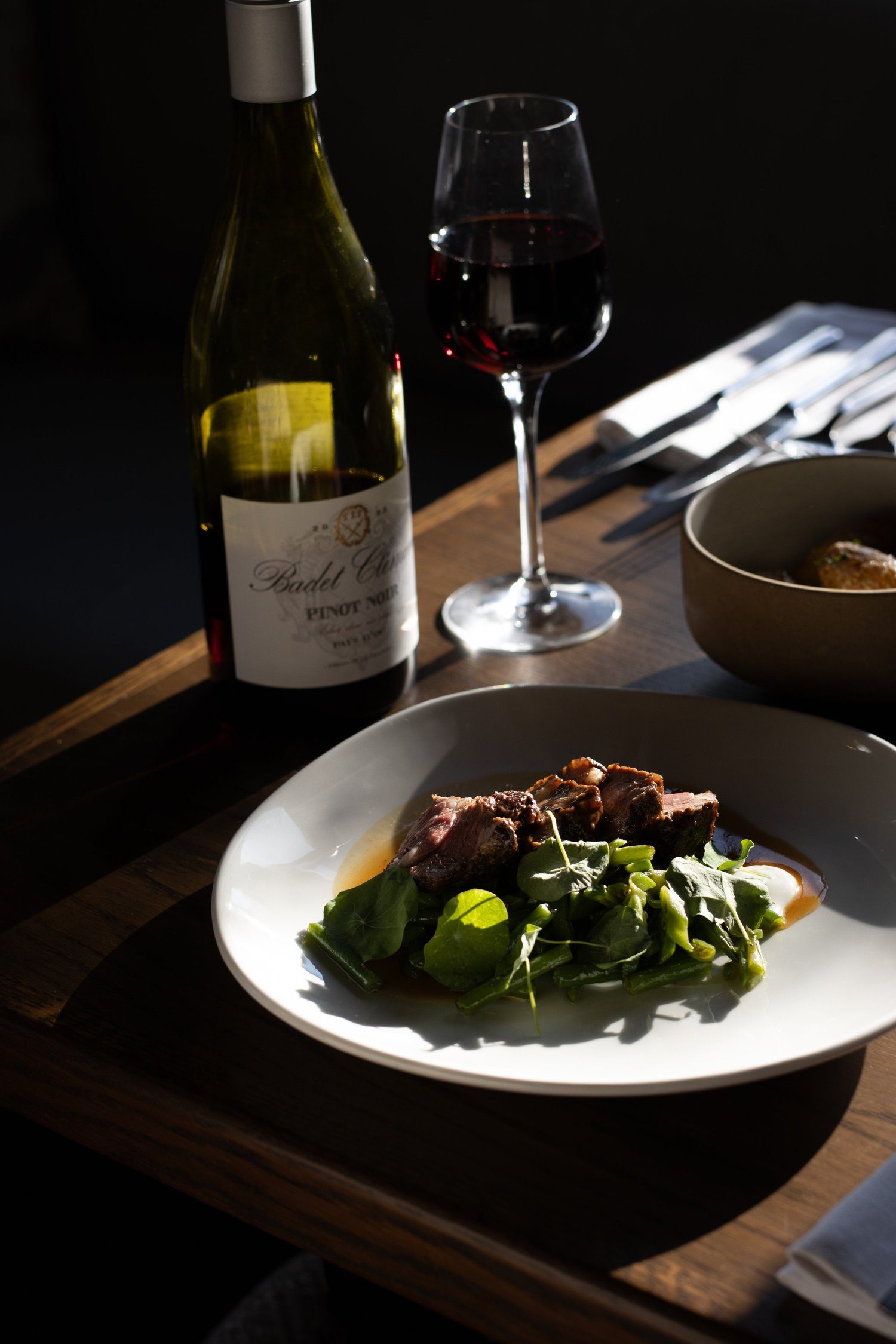 A bottle of red wine, a glass filled with red wine, a white plate with beef served with green salad, a bowl with food, and silverware on a wooden table. The Lamb Inn.