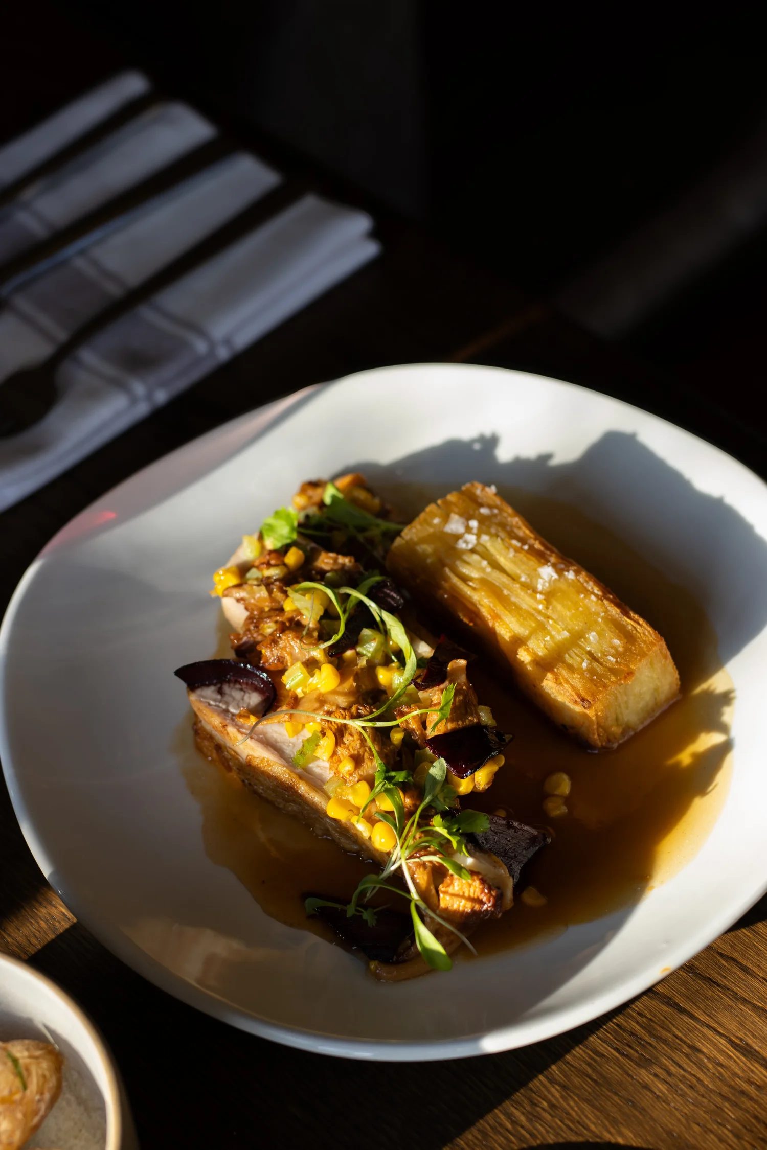 A white bowl with a piece of roasted pork topped with yellow corn kernels, black beans, and green microgreens, and a roasted potato on the side, all in a light sauce. The bowl is on a wooden table with another dish partially visible in the corner.