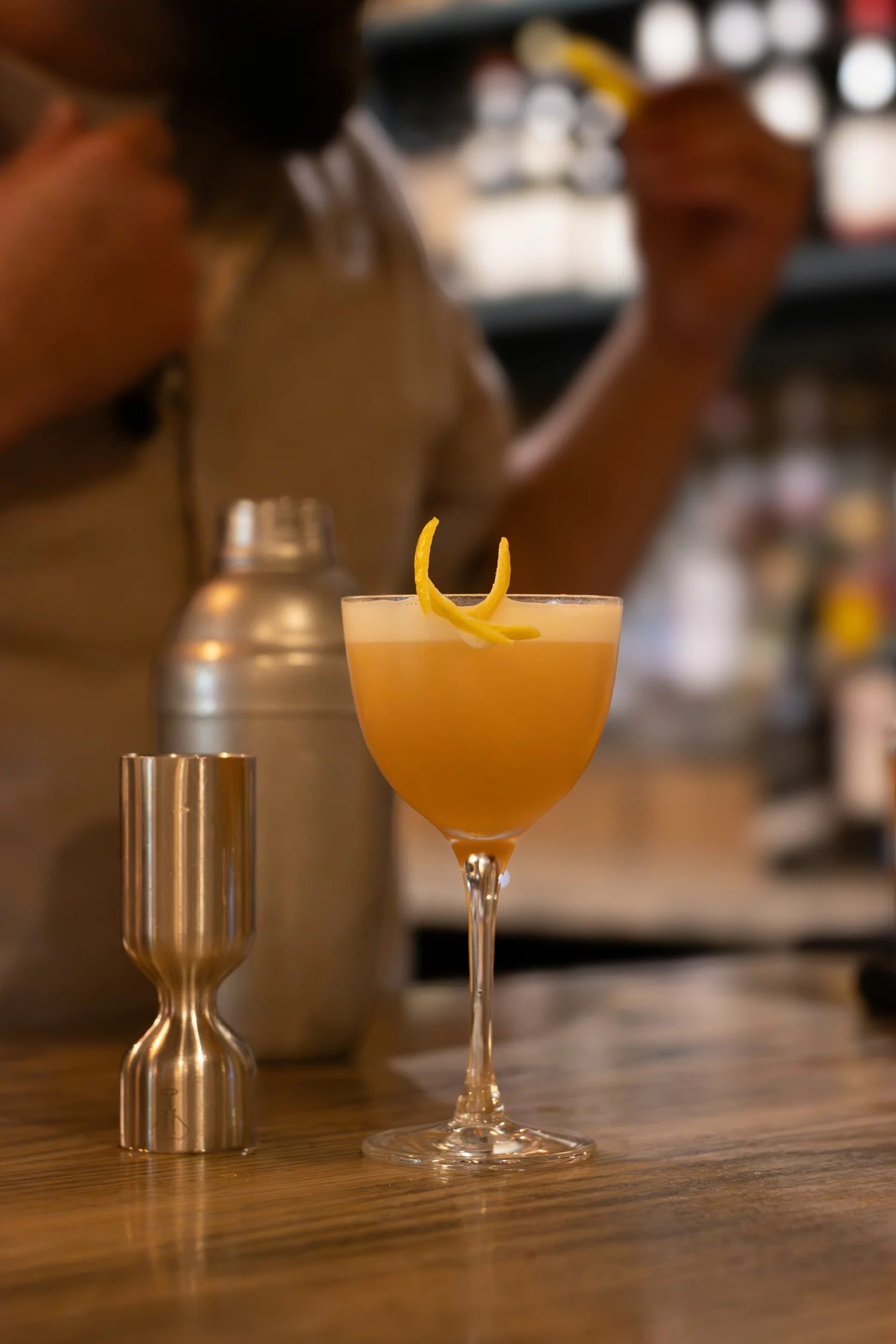 Close-up of a cocktail glass with a lemon twist garnish on a wooden bar counter, with blurred bartender in the background. The Lamb Inn.