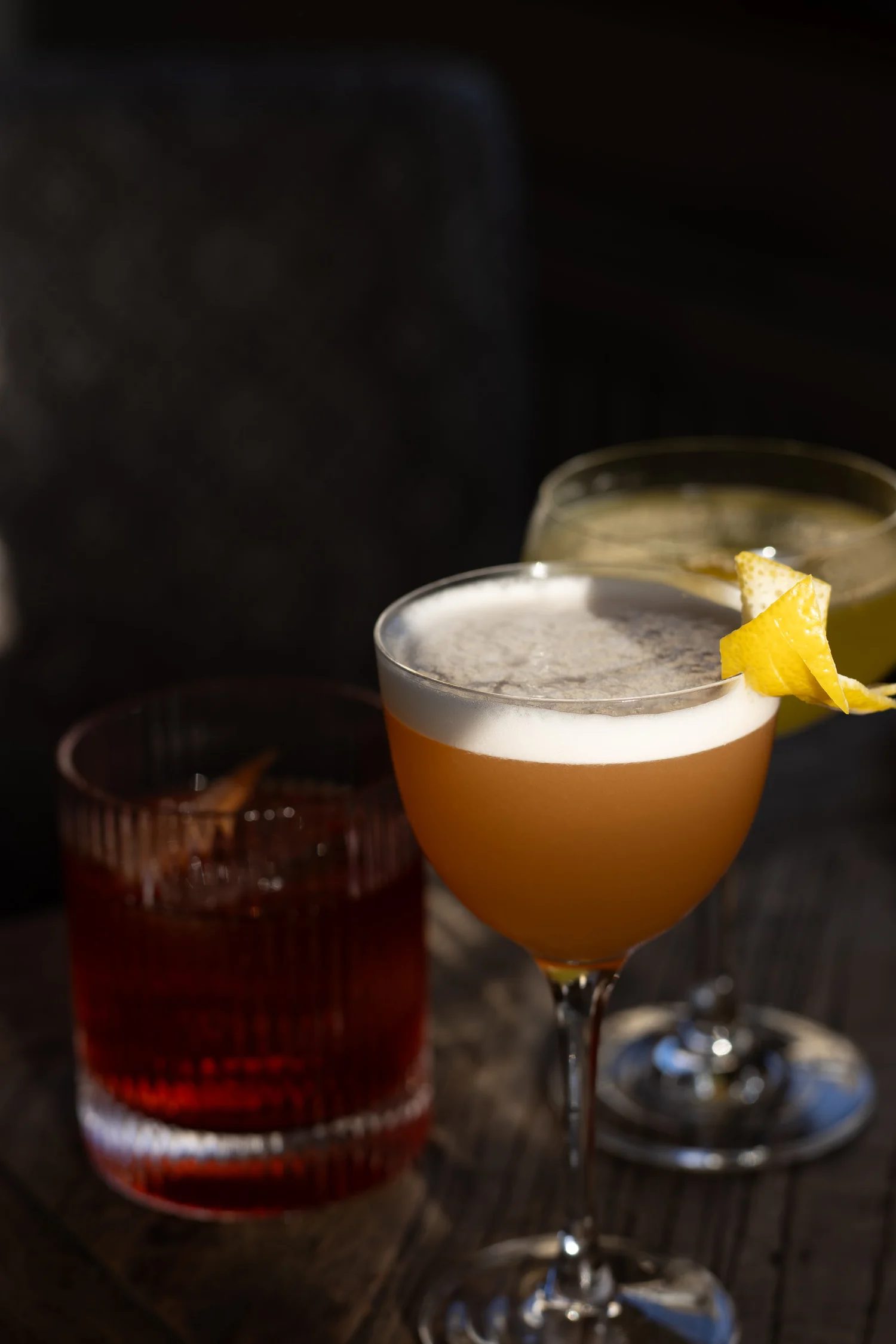 Two cocktails in martini glasses with lemon and orange slices garnishes, with a glass of amber-colored beverage in the background, on a dark table. The Lamb Inn.
