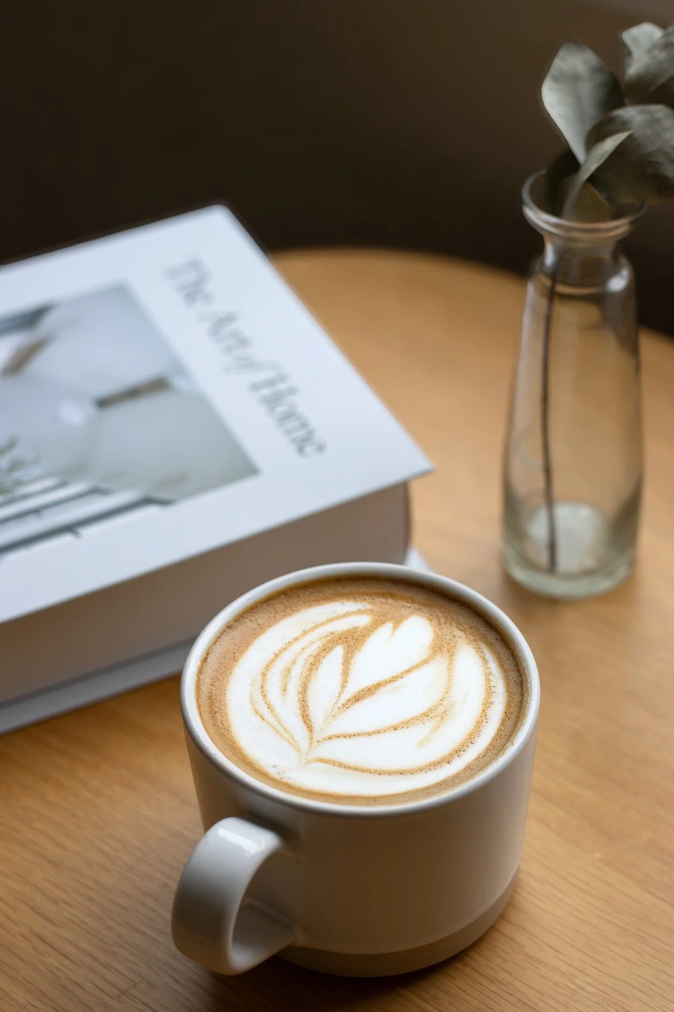 A white ceramic mug of coffee with latte art on top, placed on a wooden table. A glass vase with dried leaves and a white box with partly visible text are in the background. The New England Coffee House.