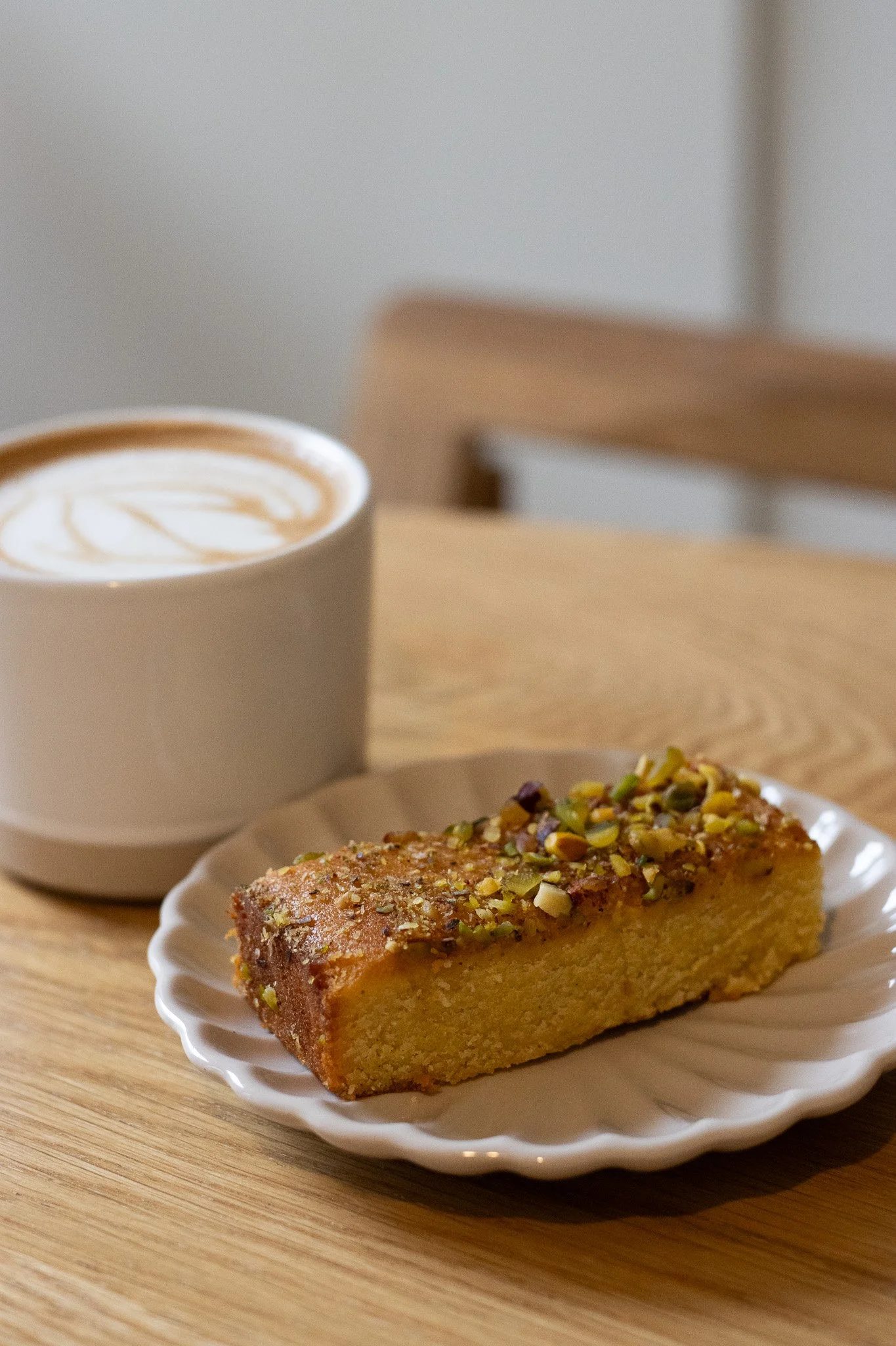 A slice of cake with chopped pistachios on top on a white plate and a cup of latte with a caramel swirl on foam in the background on a wooden table. The New England Coffee House.