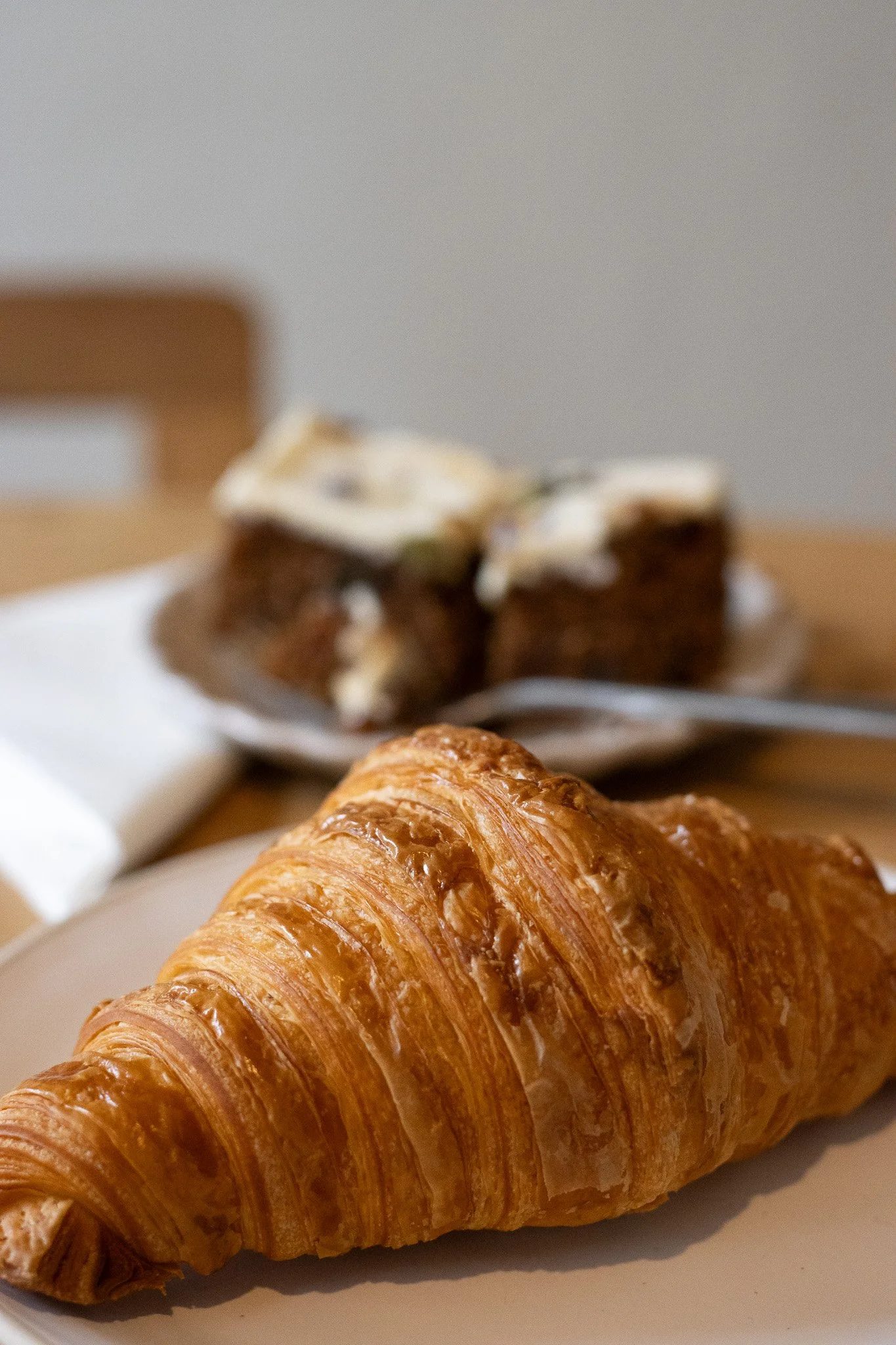 Close-up of a golden-brown, flaky croissant on a white plate, with blurred baked goods and a fork in the background. The New England Coffee House.
