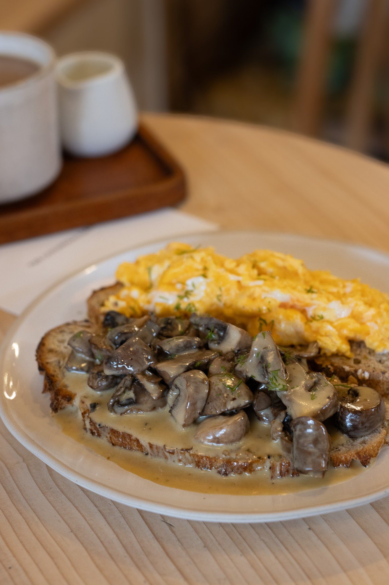 Close-up of avocado toast topped with sautéed mushrooms, scrambled eggs, and shredded cheese, served on a white plate on a wooden table. The Cotswold Farm.