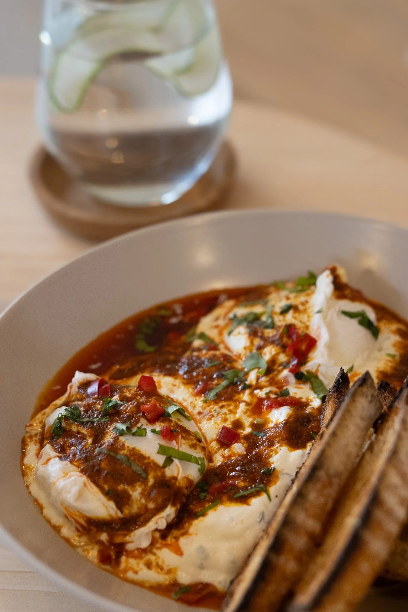 A bowl of chilaquiles topped with cream, chopped herbs, and red sauce, with a side of grilled bread and a glass of water with lime slices on a coaster. The Cotswold Farm.