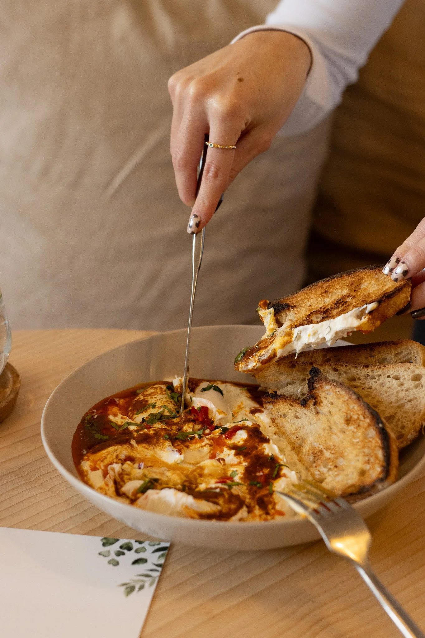 A hand with a gold ring and painted nails is holding a slice of garlic bread. The garlic bread is next to a bowl of baked Italian pizza with cheese, tomato sauce, basil, and other toppings. The bowl is on a wooden table, with a white paper napkin und