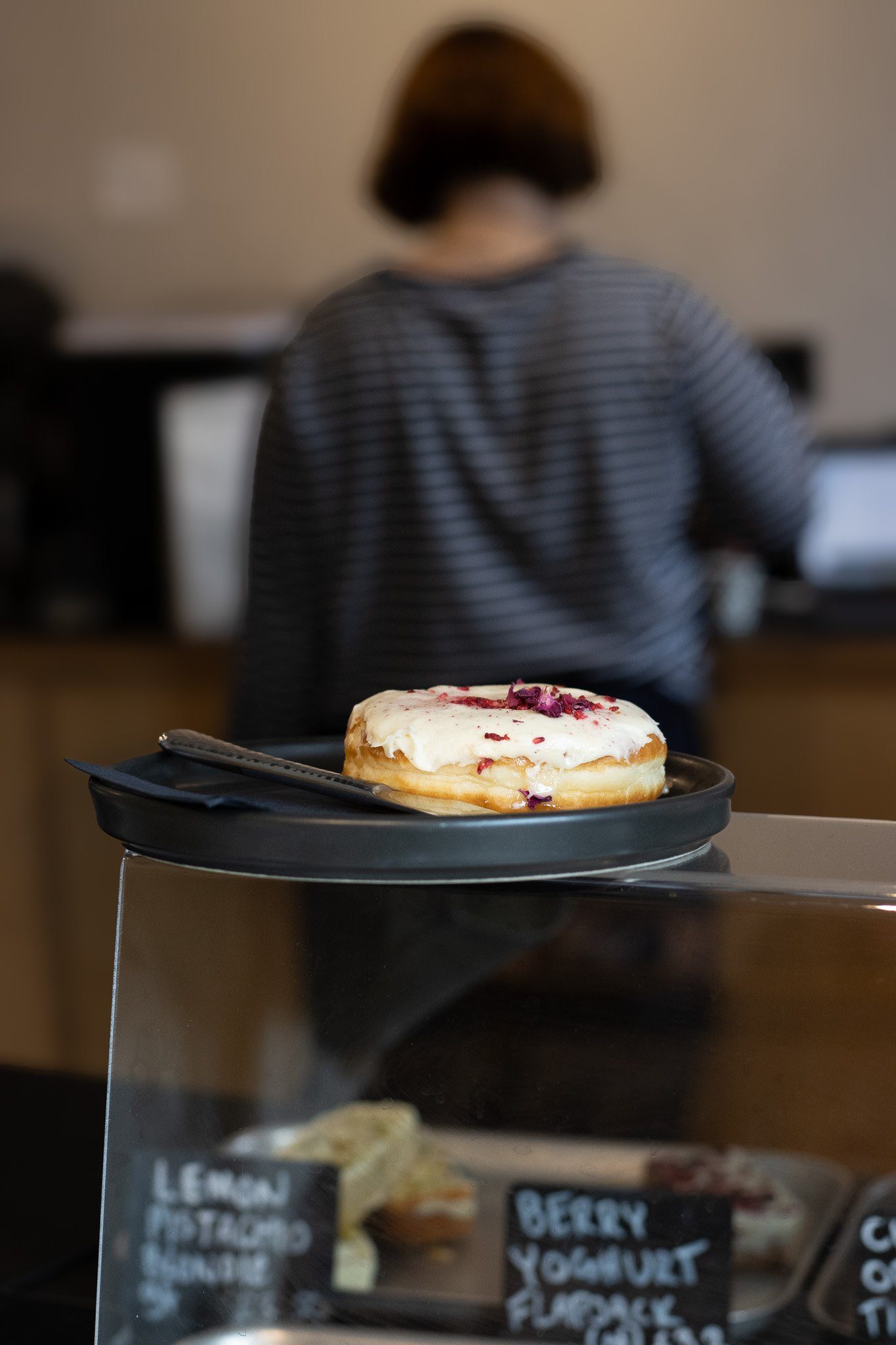 A donut with white icing and purple floral decoration, placed on a black tray with a fork, in front of a blurred person wearing a striped shirt. The Scandinavian Coffee Pod.