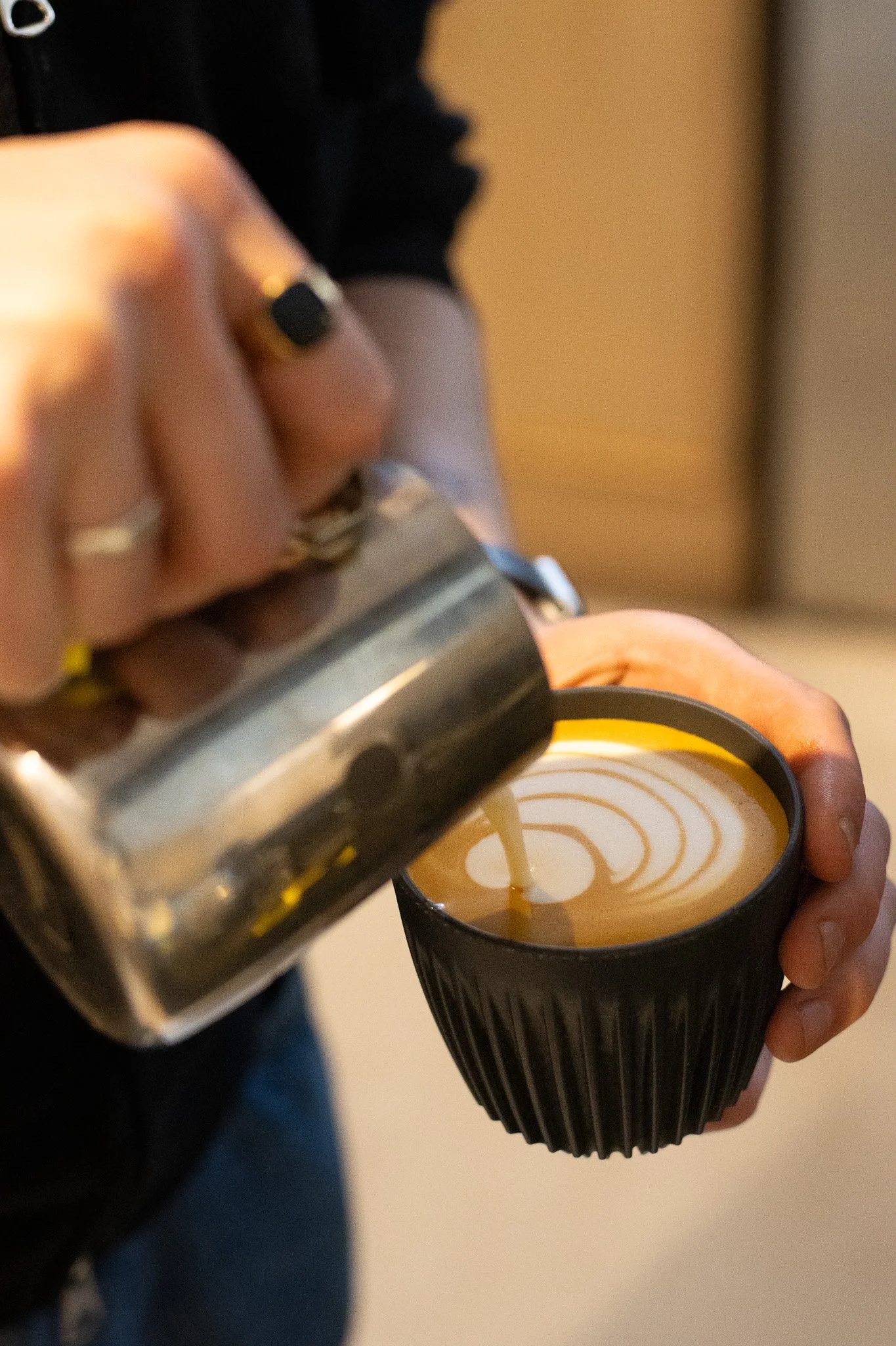 Person pouring caramel sauce from a bottle into a black cup of coffee with latte art. The Scandinavian Coffee Pod.