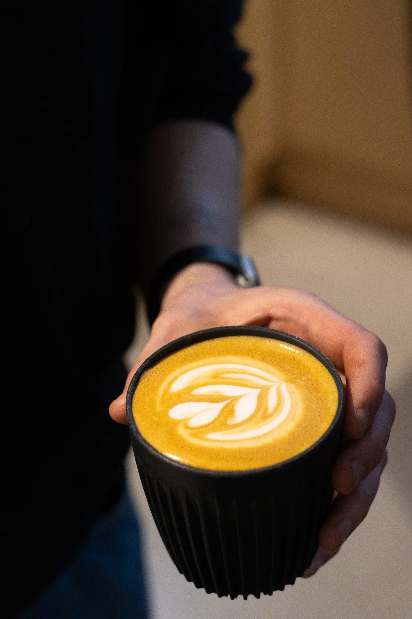 A person holding a black cup of coffee or latte with latte art in the shape of a leaf. The Scandinavian Coffee Pod.