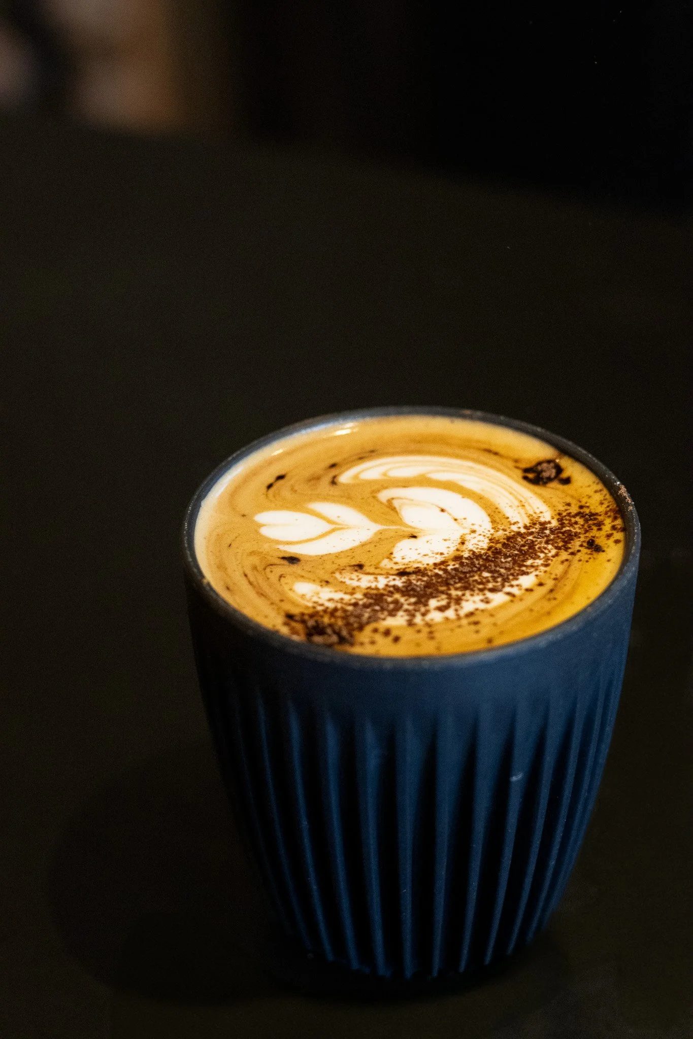 A cup of coffee with latte art and cocoa powder sprinkled on top, placed on a black surface. The Scandinavian Coffee Pod.