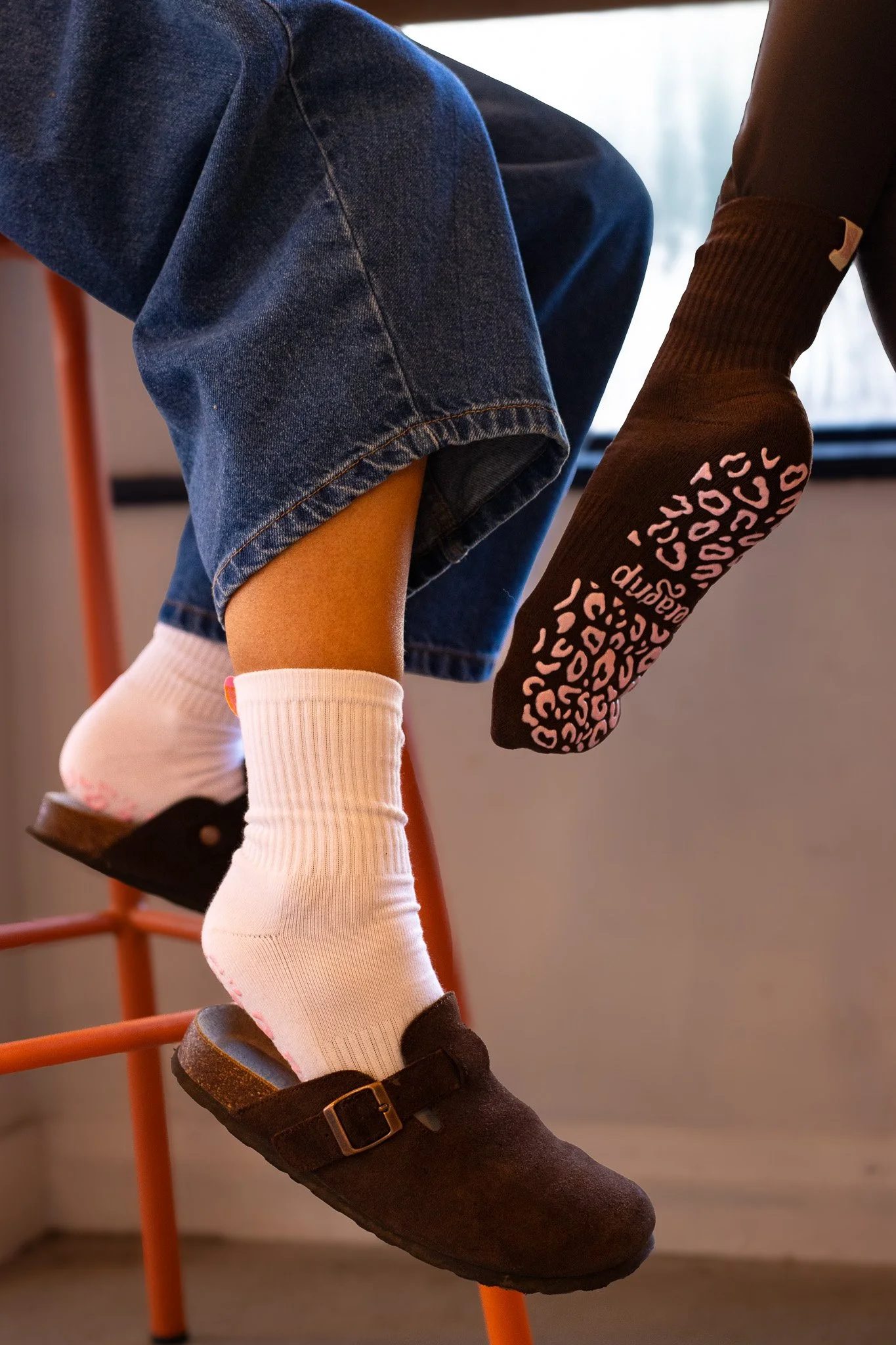 Close-up of a person wearing dark brown socks with pink text, white knee-high socks, black shoes with a buckle, denim shorts, and a black sweater, sitting on an orange chair. GetAGrip Apparel.
