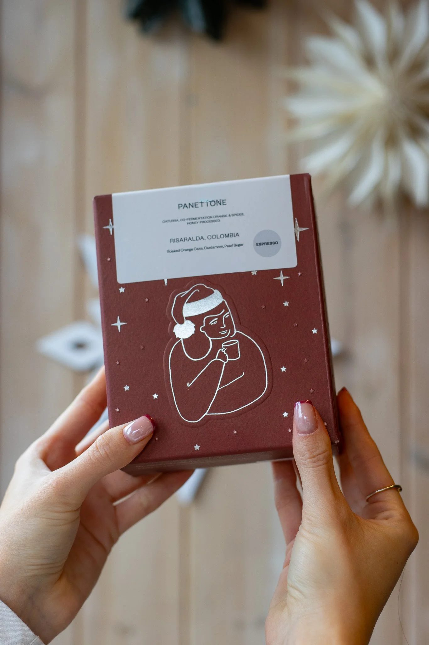 Person holding a red package with white illustration of a woman wearing a Santa hat and headphones, snowflakes and stars around, text about a Colombian orange cake flavor. The Scandinavian Coffee Pod.