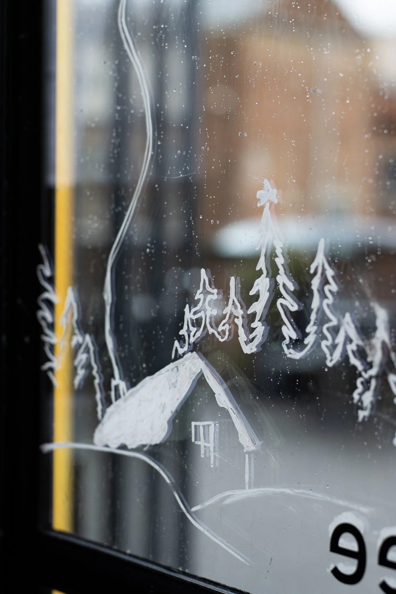 A window with doodles of trees and a mountain drawn with white marker or paint, foggy with raindrops on the glass. The Scandinavian Coffee Pod.