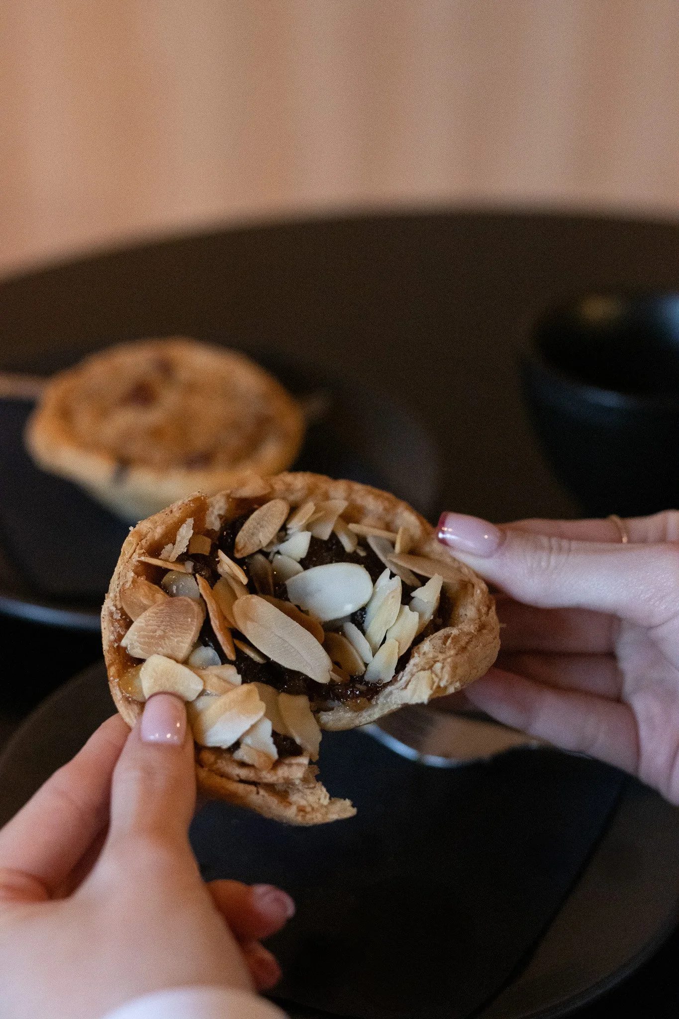 Close-up of a person holding a cookie with chocolate, sliced almonds, and a cream filling, while another cookie rests on a black plate in the background. The Scandinavian Coffee Pod.