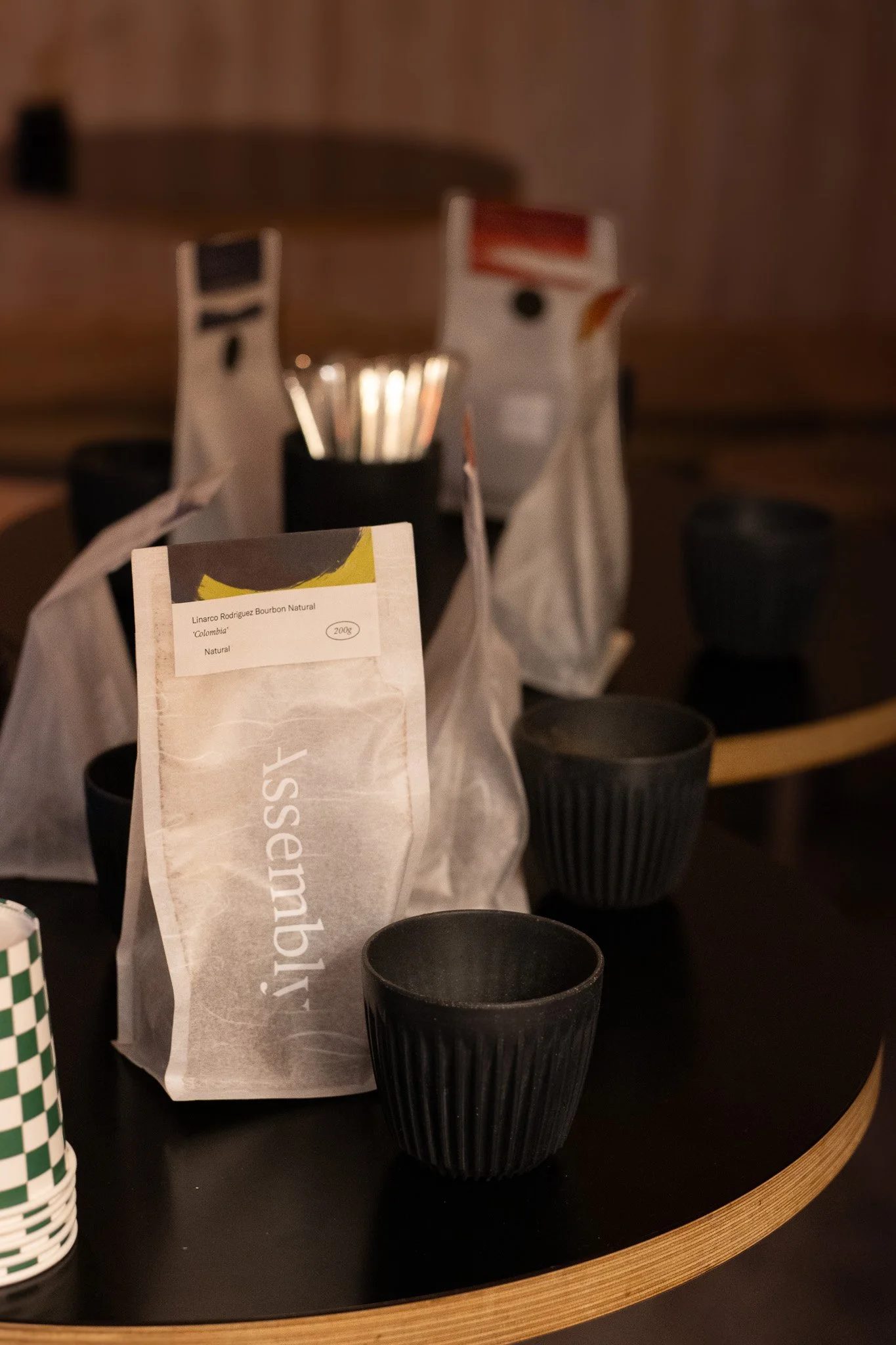 Close-up of a coffee table with black cups, a paper bag of coffee beans labeled 'Linarco Rodriguez Bourbon Natural Colombia', and a white paper bag. In the background, there are brown paper bags, a metallic object, and a wooden tray, all under dim li