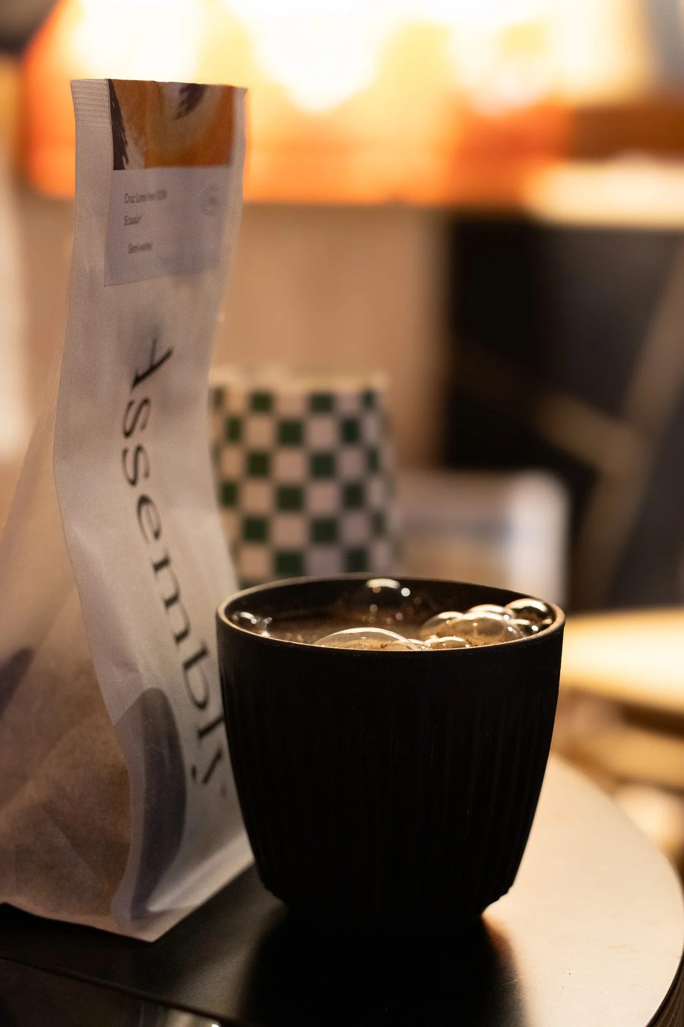 A paper bag with a coffee shop logo, a black cup with coffee and ice, and a checkered cup in the background, likely in a café setting. The Scandinavian Coffee Pod.