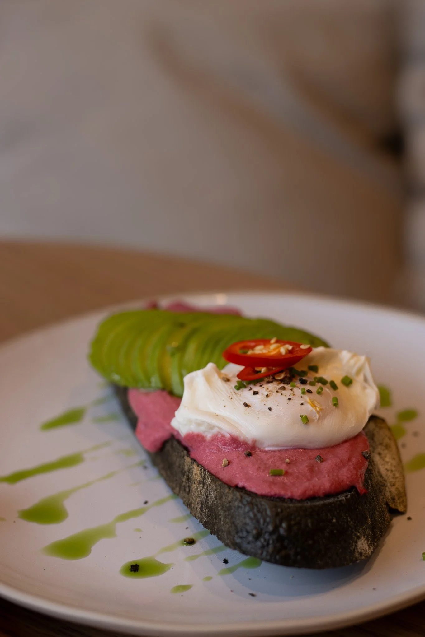 Open-faced sandwich on dark bread with pink spread, topped with a poached egg, sliced red chili, chopped chives, black pepper, and garnished with a green drizzle and black specks on a white plate. The Cotswold Farm.