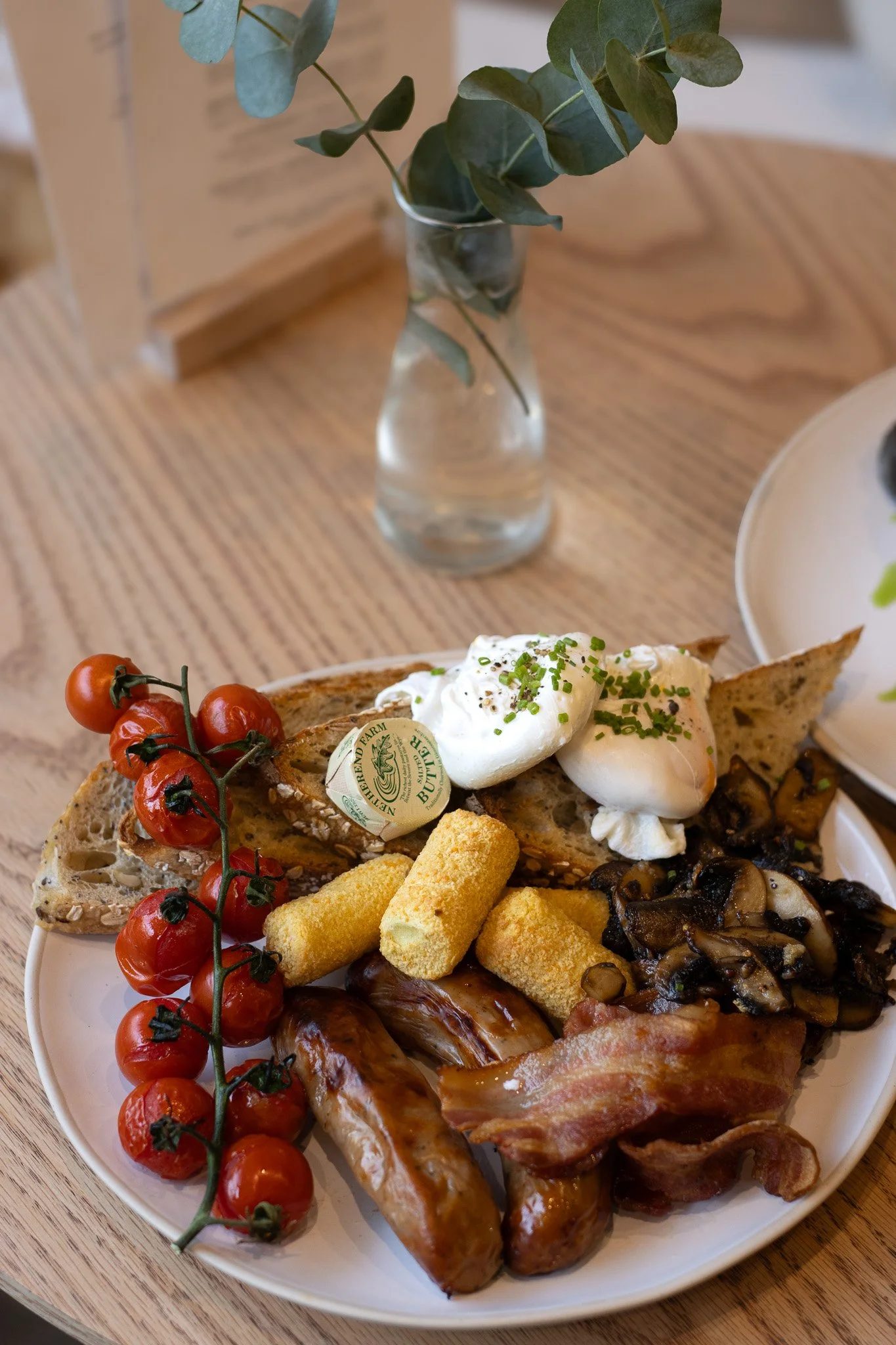 Breakfast plate with cherry tomatoes, bacon, mushrooms, eggs, potatoes, bread, and a sprig of cherry tomatoes on the side, on a white plate. The Cotswold Farm.