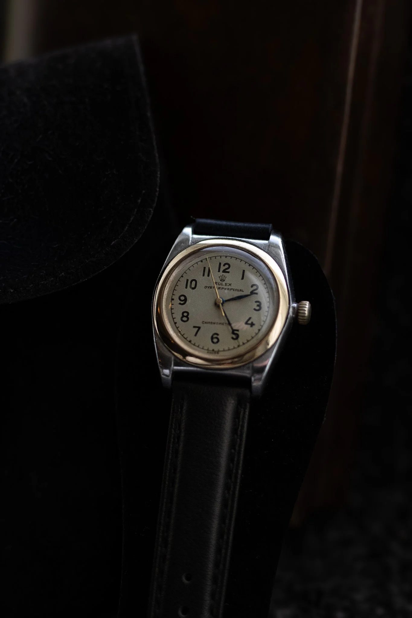 Close-up of a vintage Rolex Oyster Perpetual watch with a white dial, black numerals, and a black leather strap, resting on a black surface with a dark background. Aurmont. Watches.