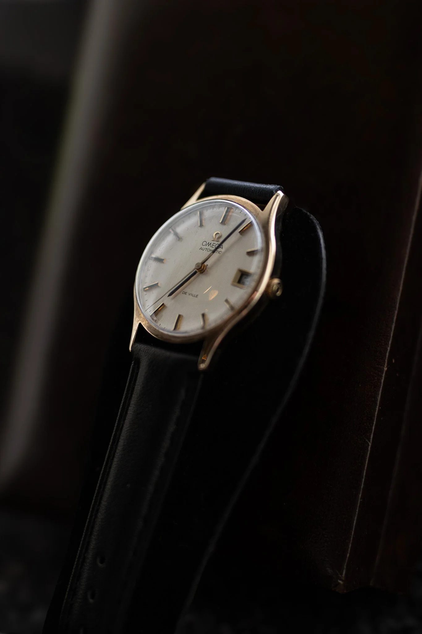 Close-up of a vintage Omega automatic wristwatch with a cream dial, gold markers and hands, and a black leather strap, resting on a dark surface. Aurmont. Watches.