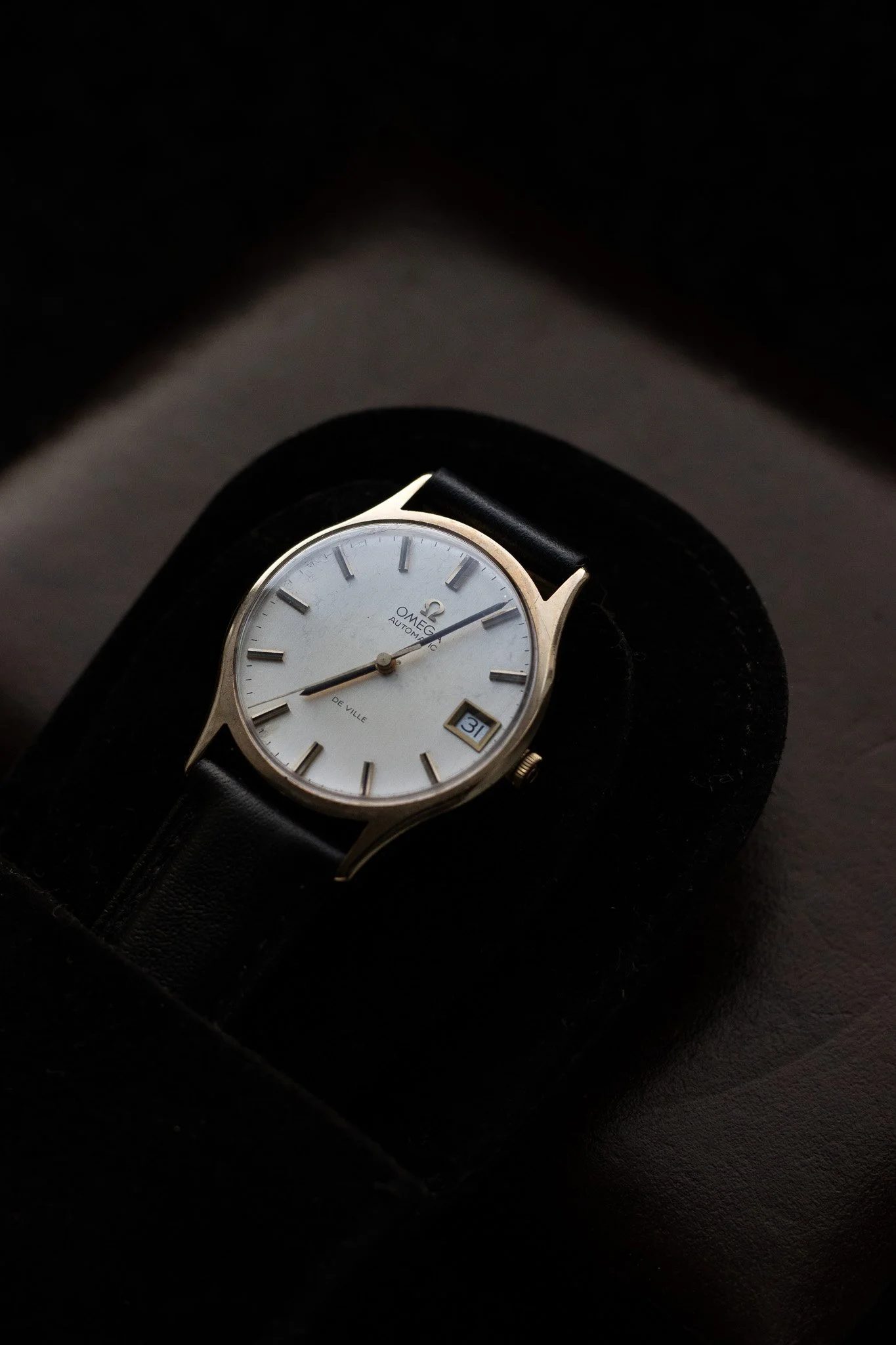 A vintage Omega De Ville wristwatch with a white dial, black leather strap, and a date window at 3 o'clock, resting on a black watch stand against a dark background. Aurmont. Watches.