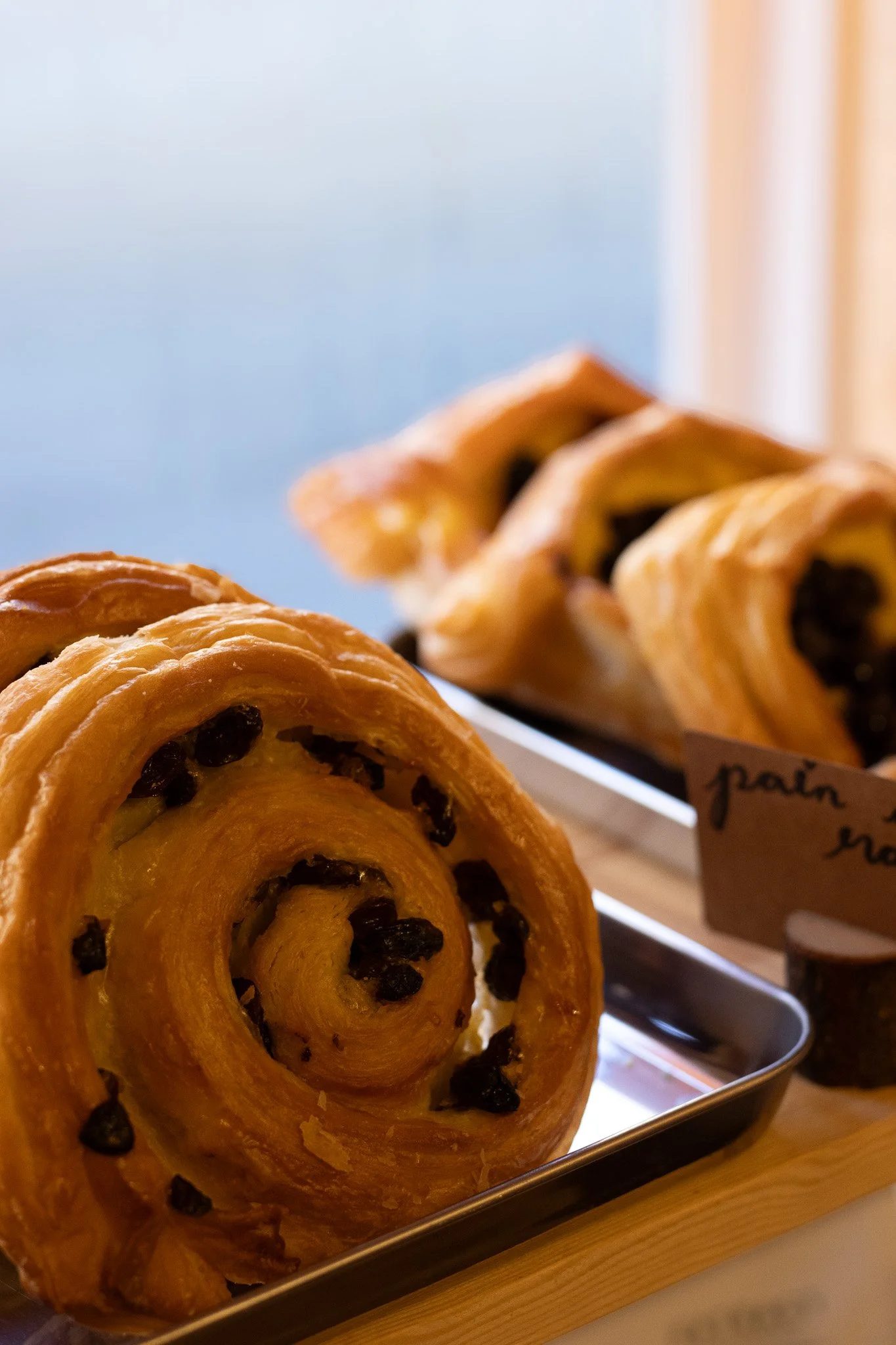 Close-up of a pastry roll with dark filling, on a metal tray, with a background of a sky and blurred interior. The New England Coffee House.