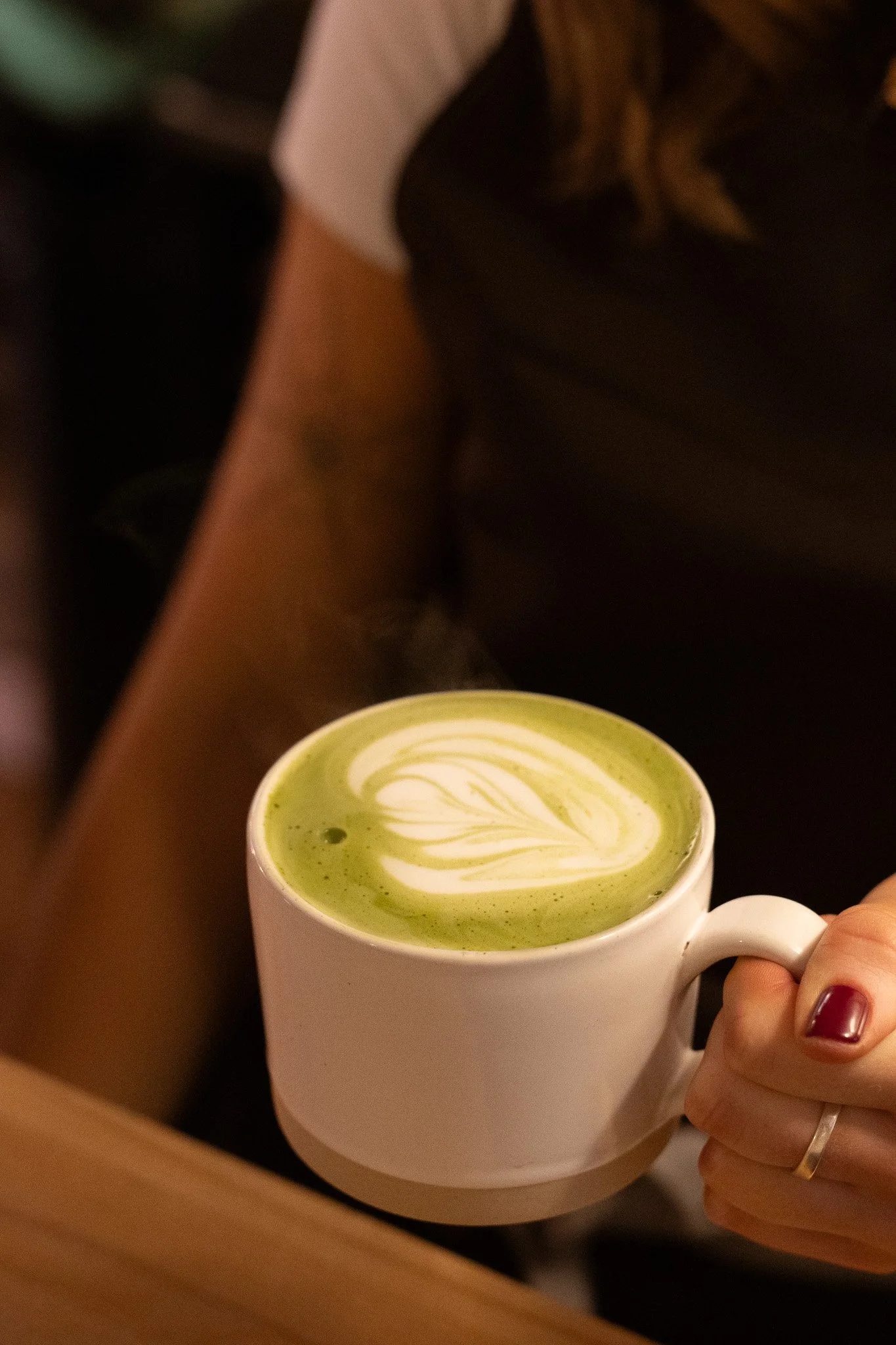 A person holding a white mug filled with green matcha latte with latte art. The New England Coffee House.