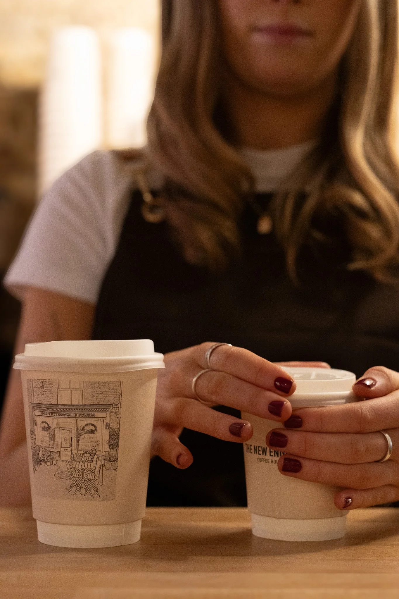 A person with long, wavy blonde hair and dark red nail polish holding two white coffee cups, with one cup on the table and the other in their hands, in a cozy indoor setting. The New England Coffee House.