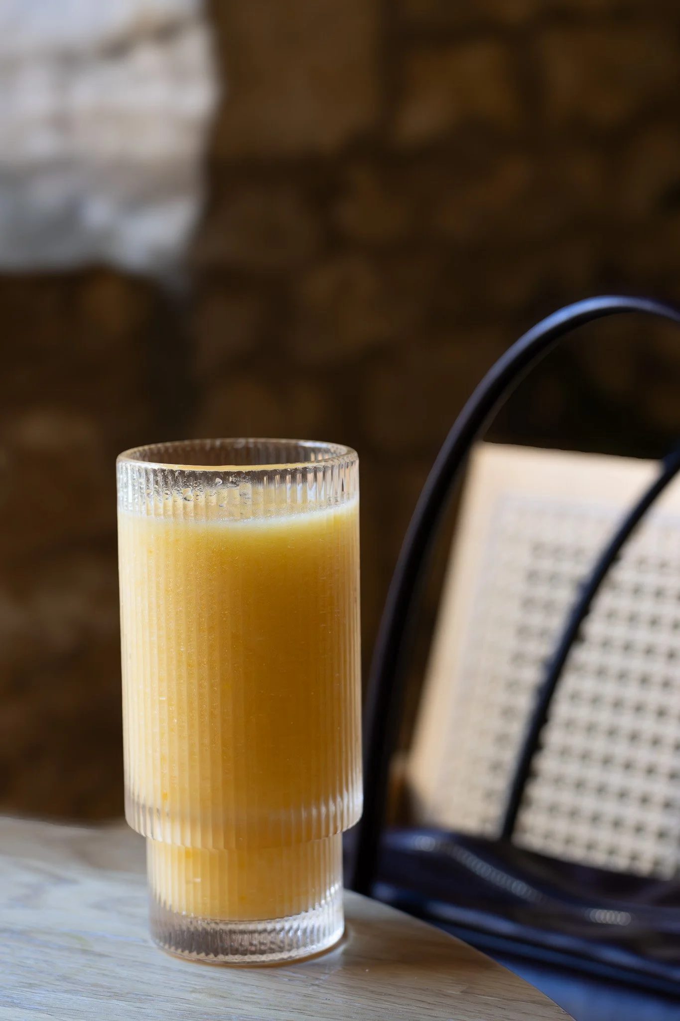 A glass filled with a yellowish liquid, placed on a wooden table, with a blurred background that includes a brick wall and a black wire. The New England Coffee House.