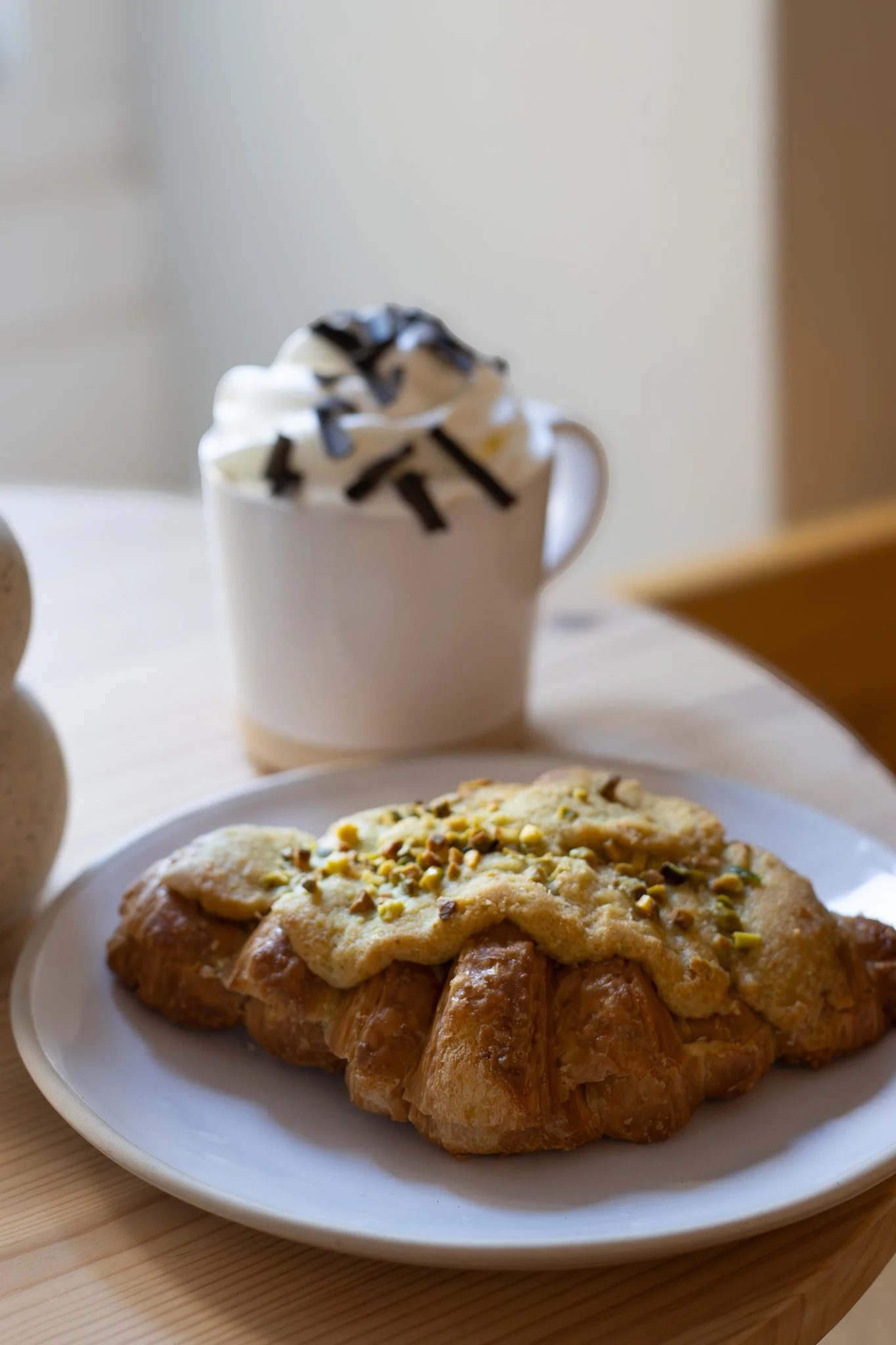 A white plate with a croissant topped with glazed cream and chopped pistachios. In the background, a cup of whipped cream with chocolate drizzle and a dessert, possibly a cookie or cake, on a white plate. The scene is set on a light wooden surface.